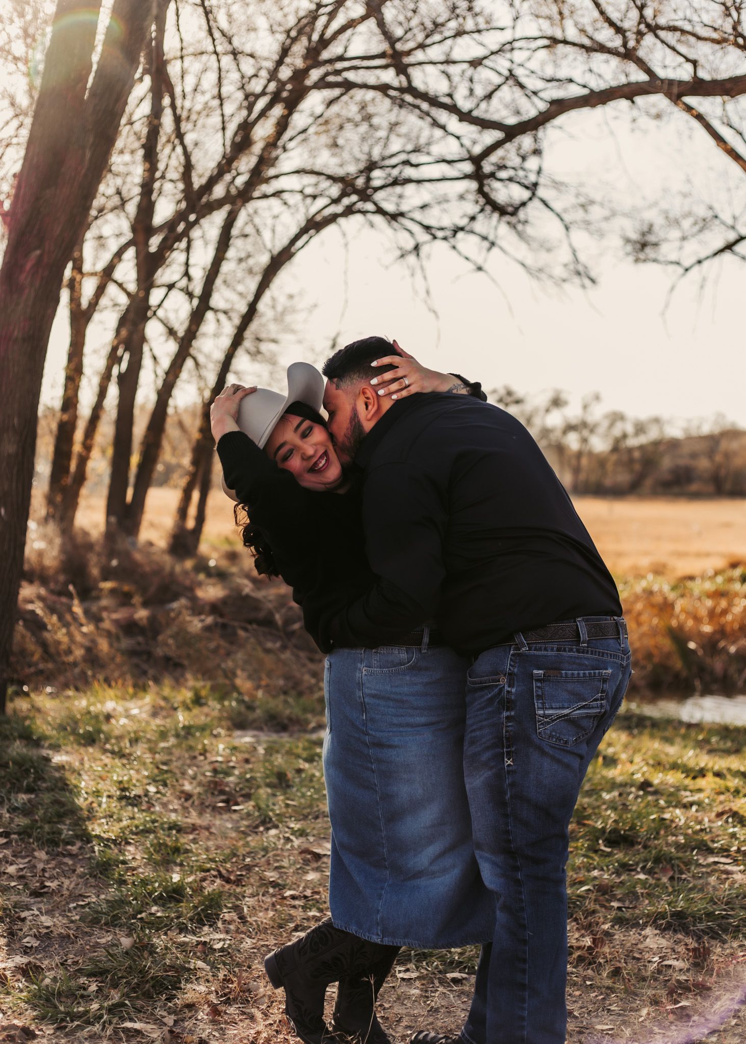 Couple sharing a tender embrace in a sunset-lit wooded area wearing casual denim outfits.