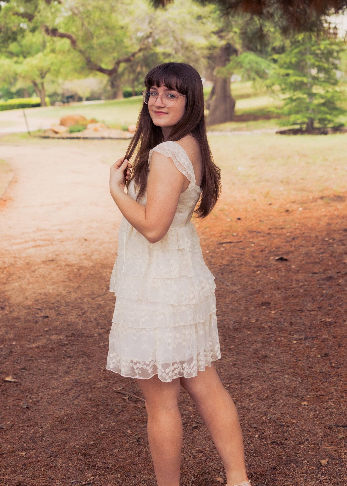 A person in a white lace dress poses outdoors in a garden setting with trees in the background.