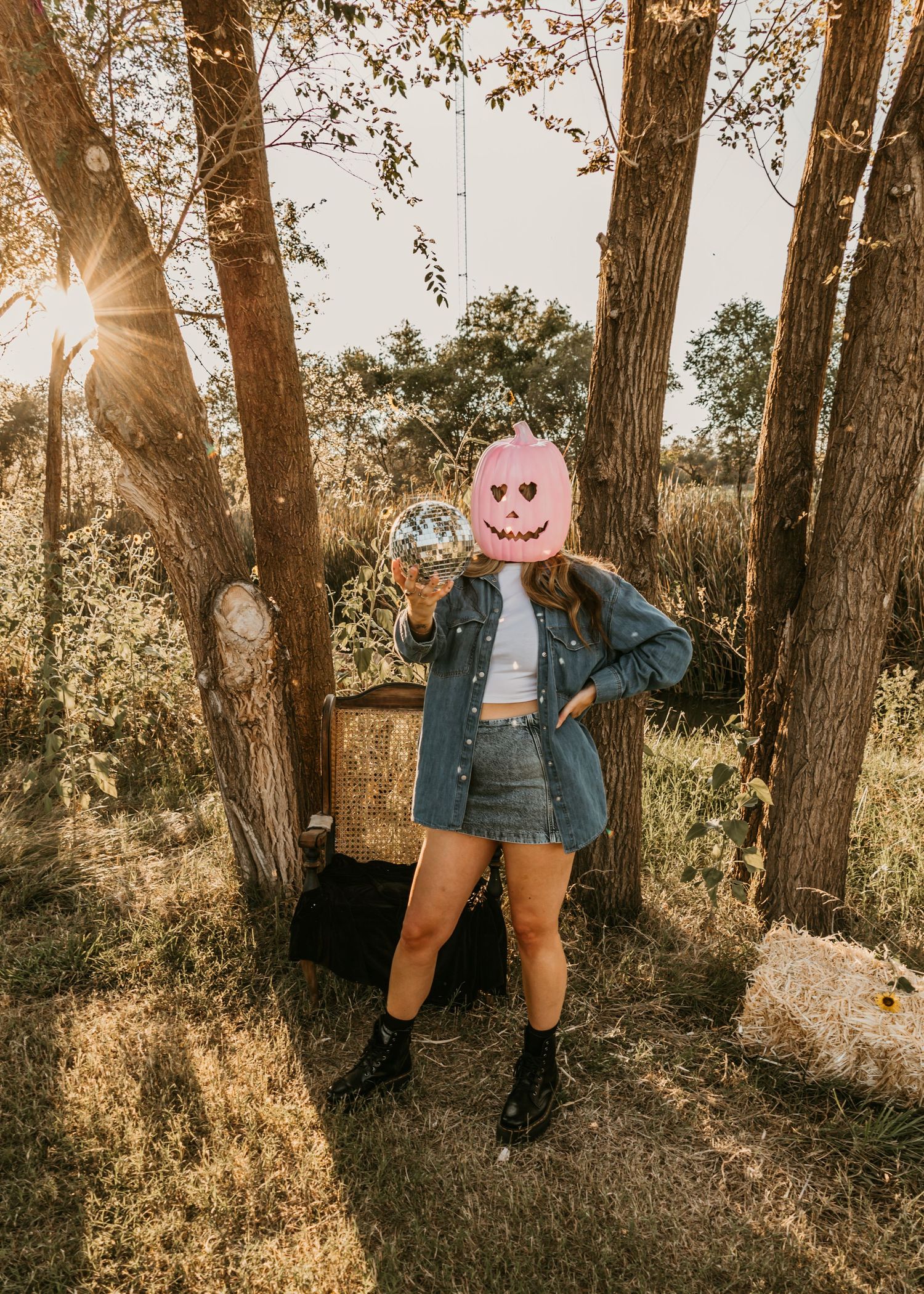 Person holding disco ball with pink pumpkin mask, wearing denim outfit and boots, standing among trees in golden sunlight.