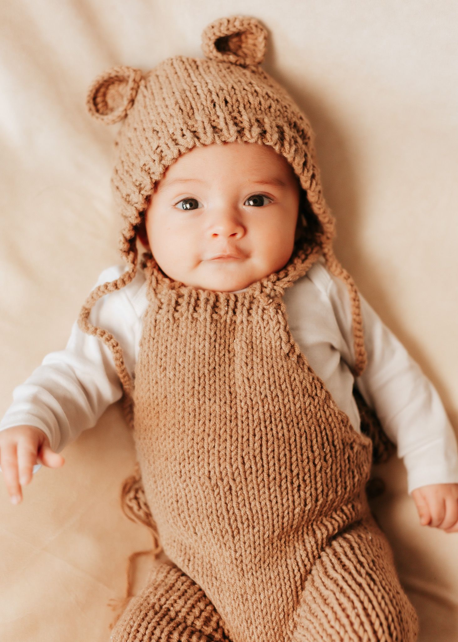 Baby wearing knitted brown bear outfit with ears lying on cream colored background.