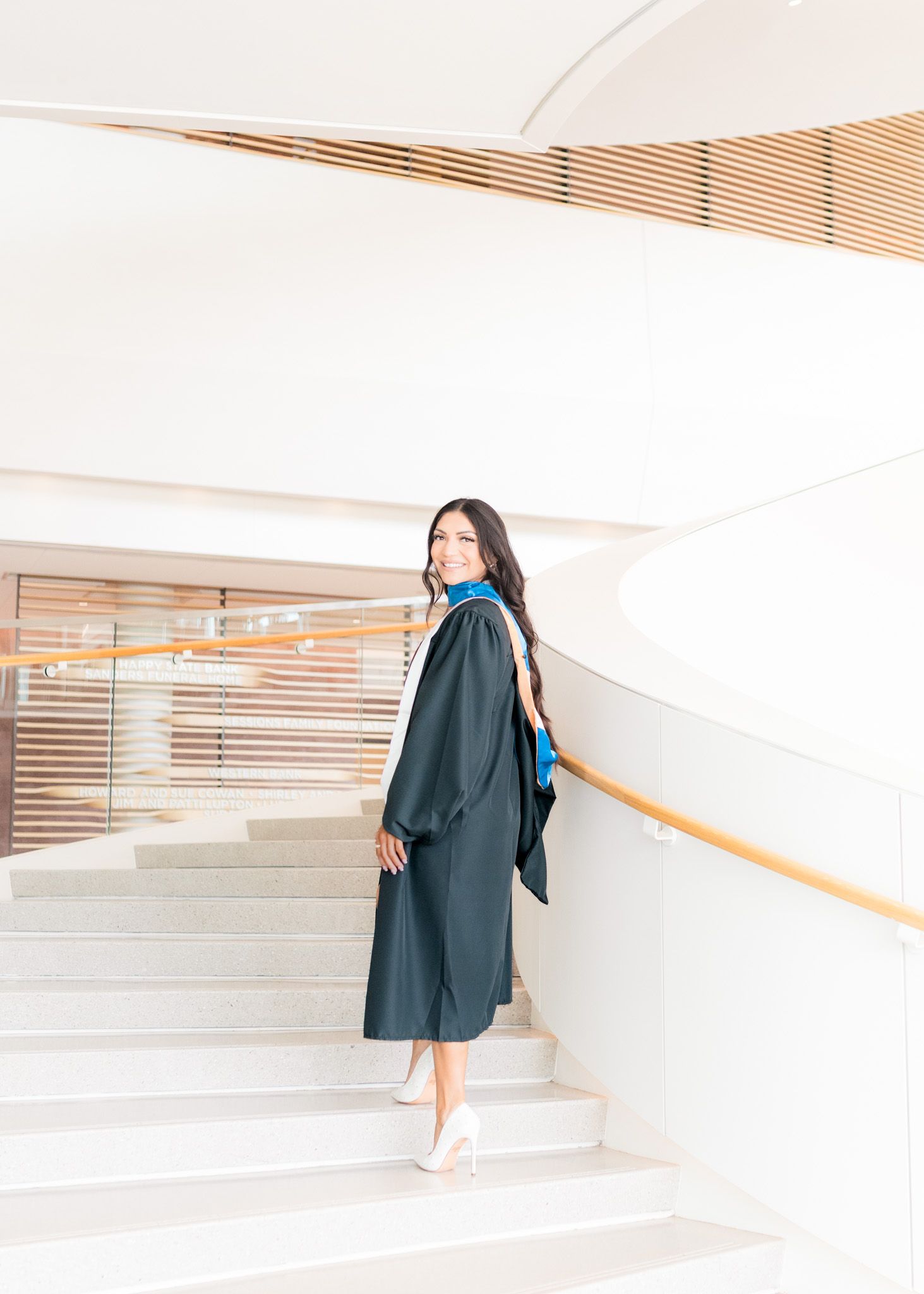 Graduate in dark academic regalia poses on modern white staircase with handrail.