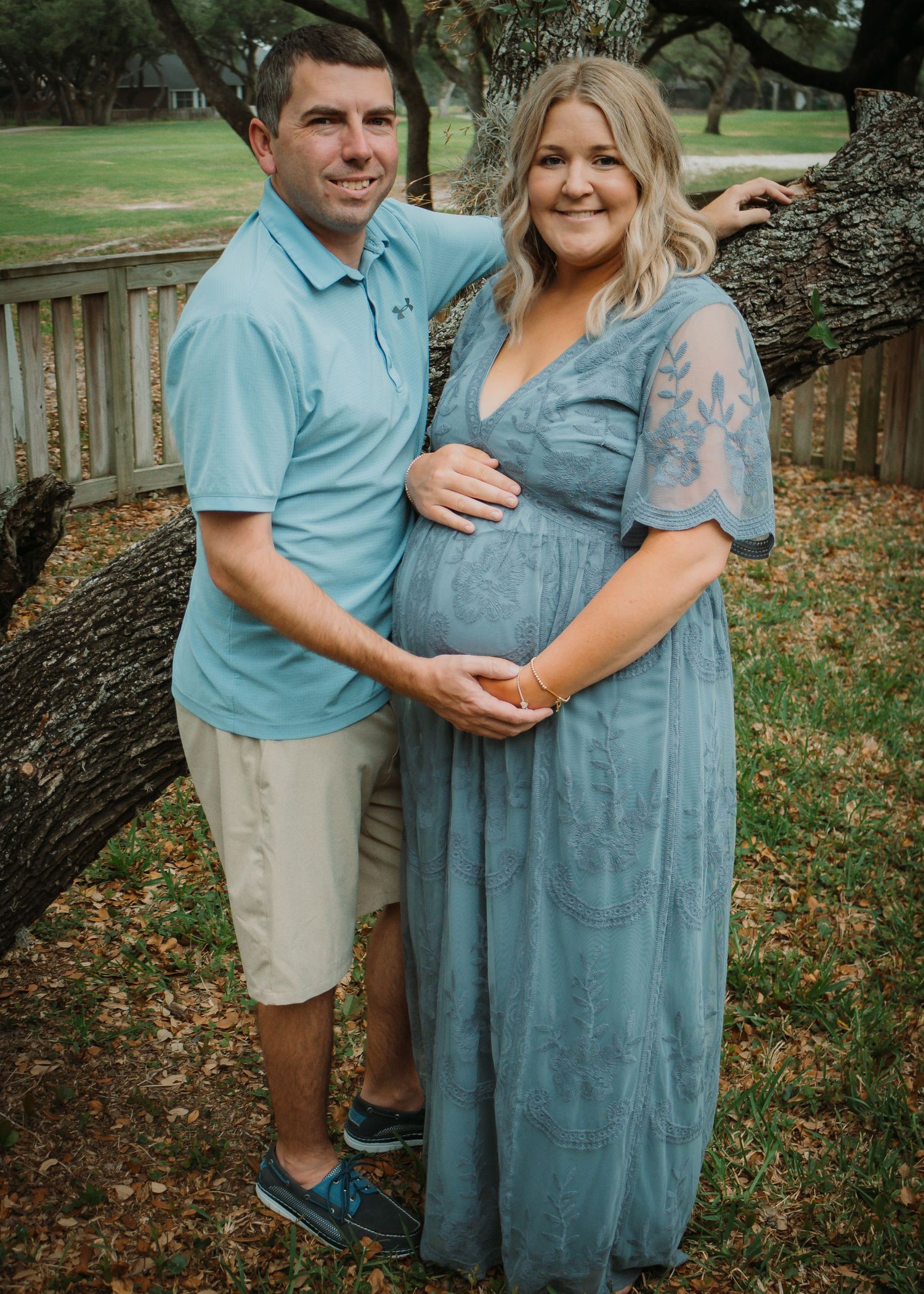 Outdoor maternity photo of couple in blue and grey attire by wooden fence.