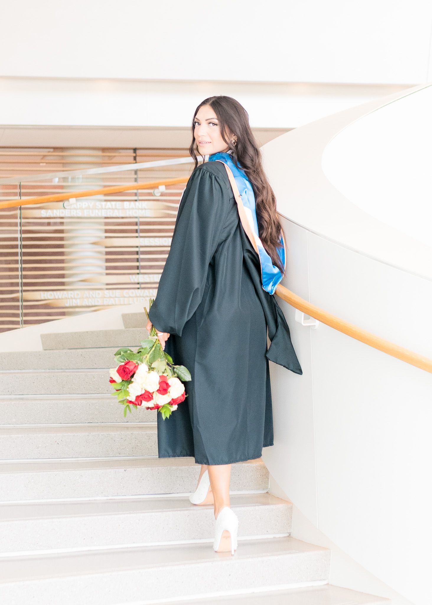 Graduate in black gown holds flowers while ascending stairs in cap and gown.