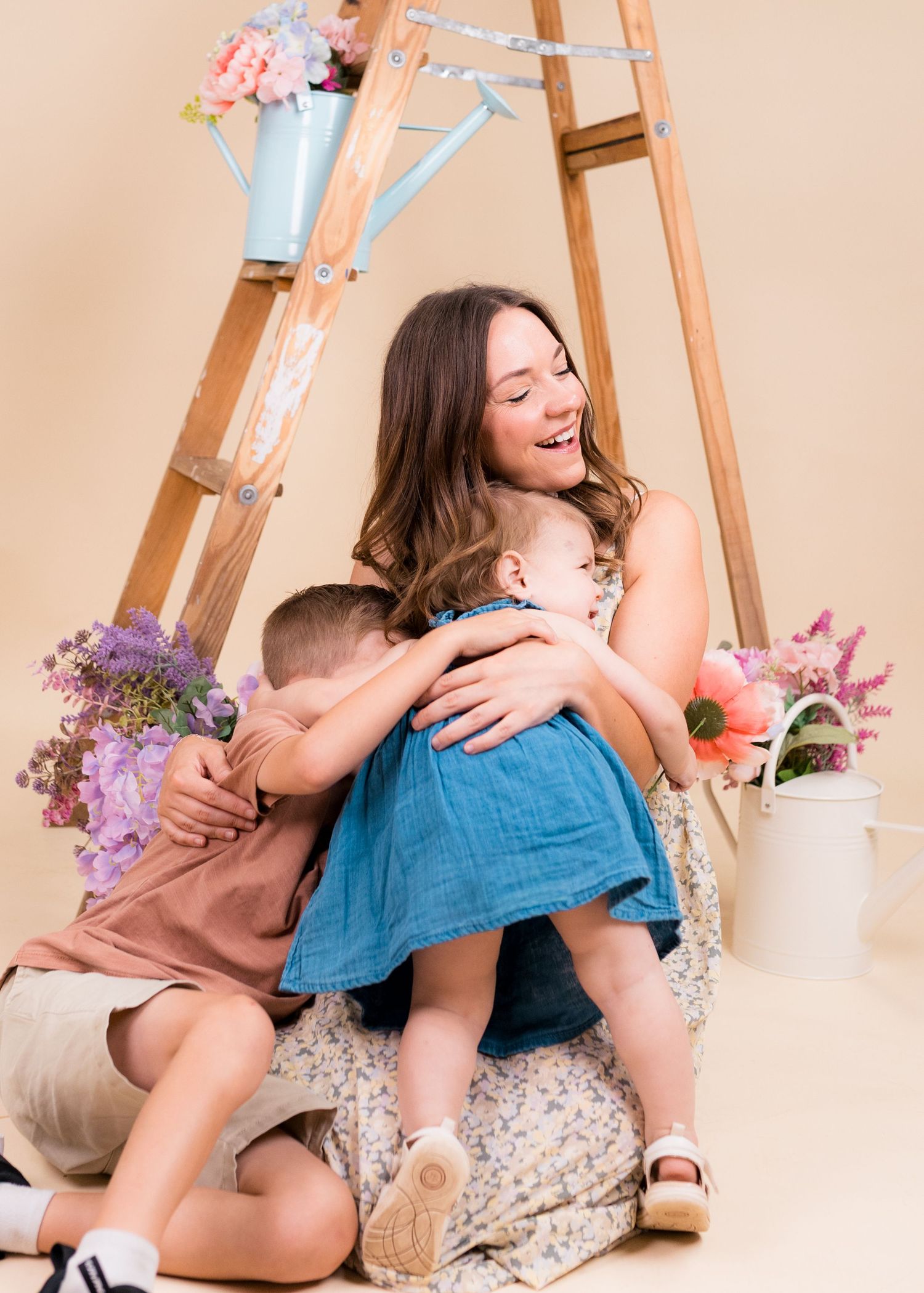 A laughing mother hugs her two young children in a bright studio with flowers and a wooden ladder backdrop.