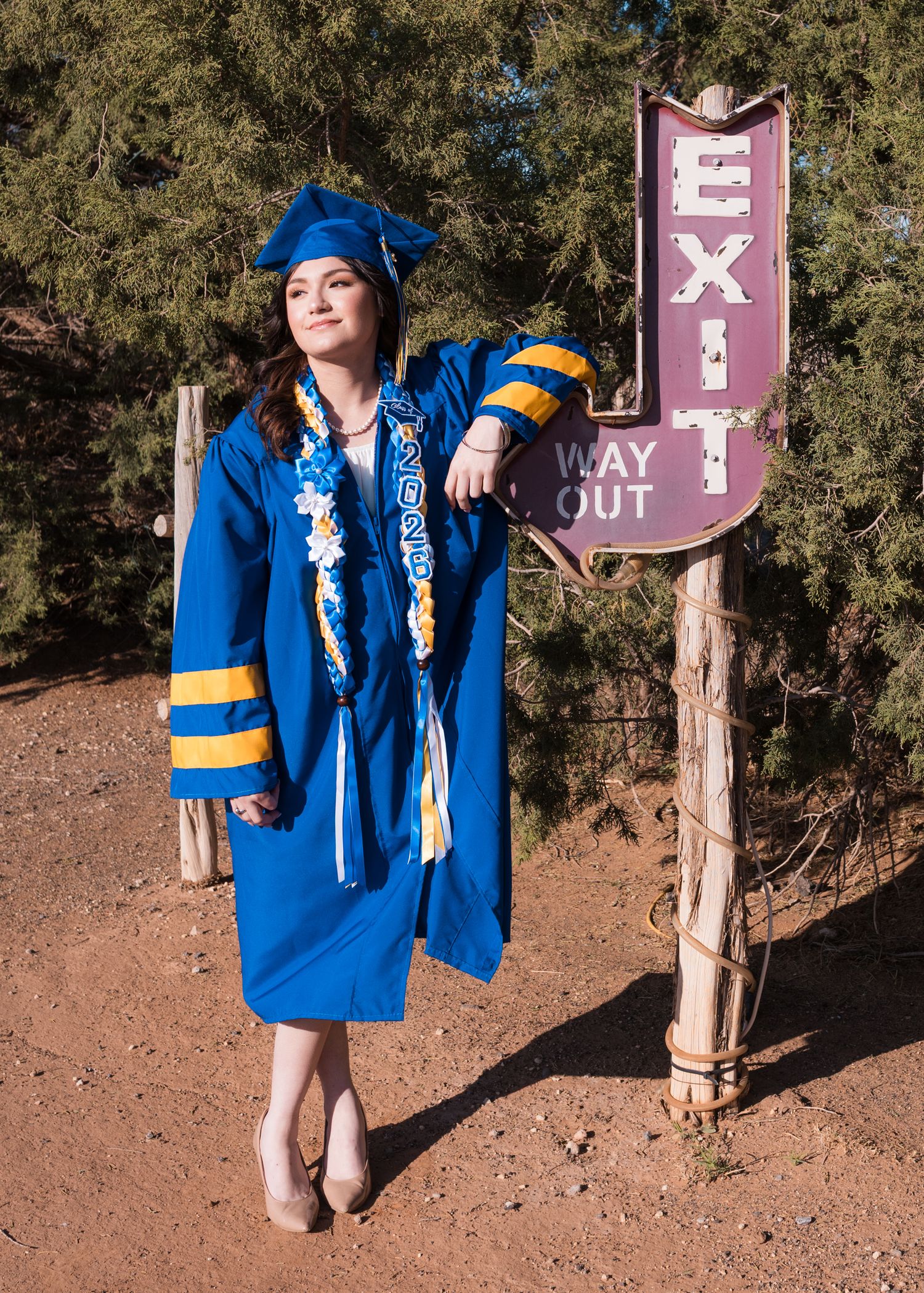 Graduate in blue cap and gown poses beside an Exit sign outdoors, wearing a 2023 sash and floral lei.