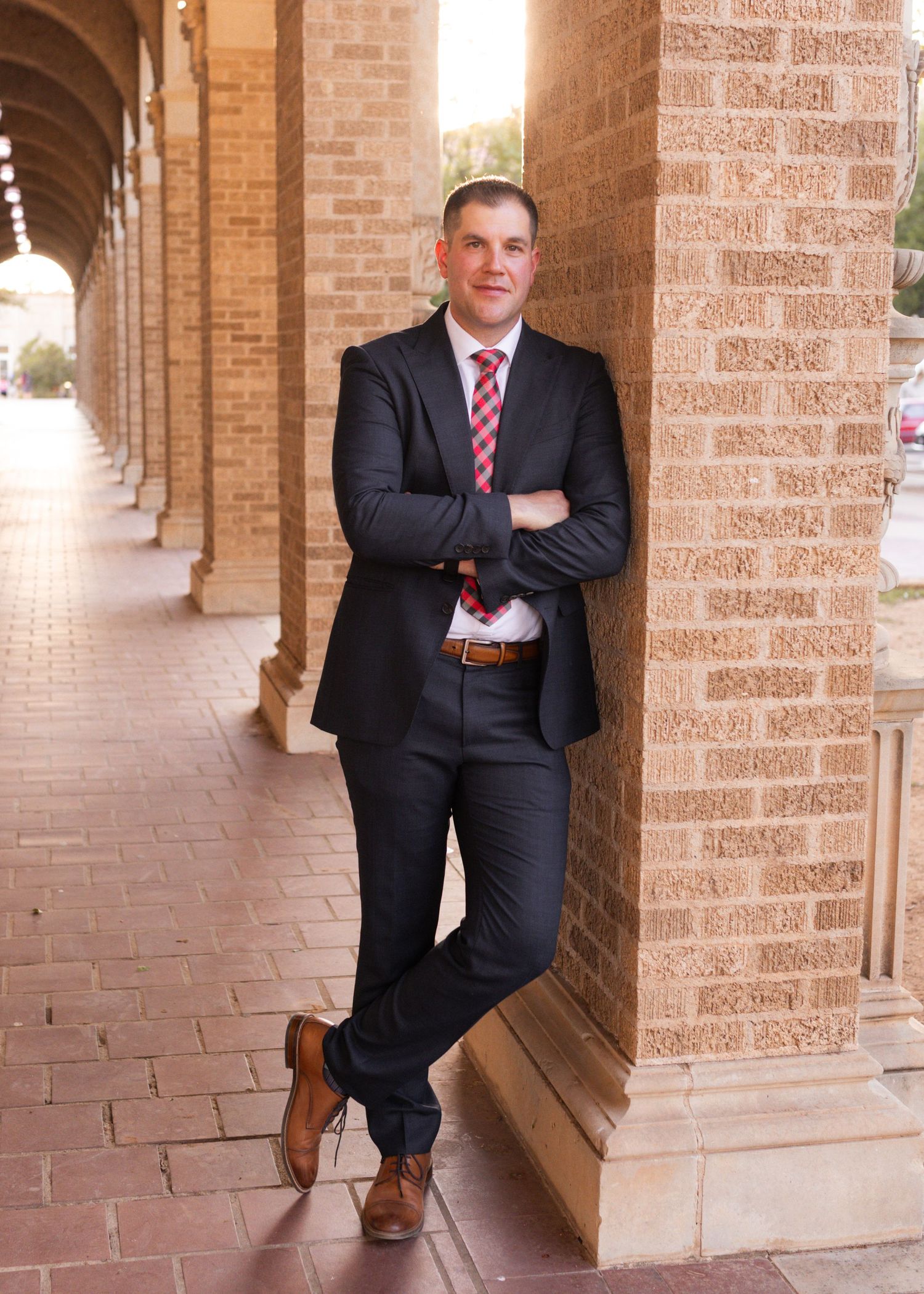 Professional man in dark suit and red tie leaning against brick columns in an elegant outdoor corridor.