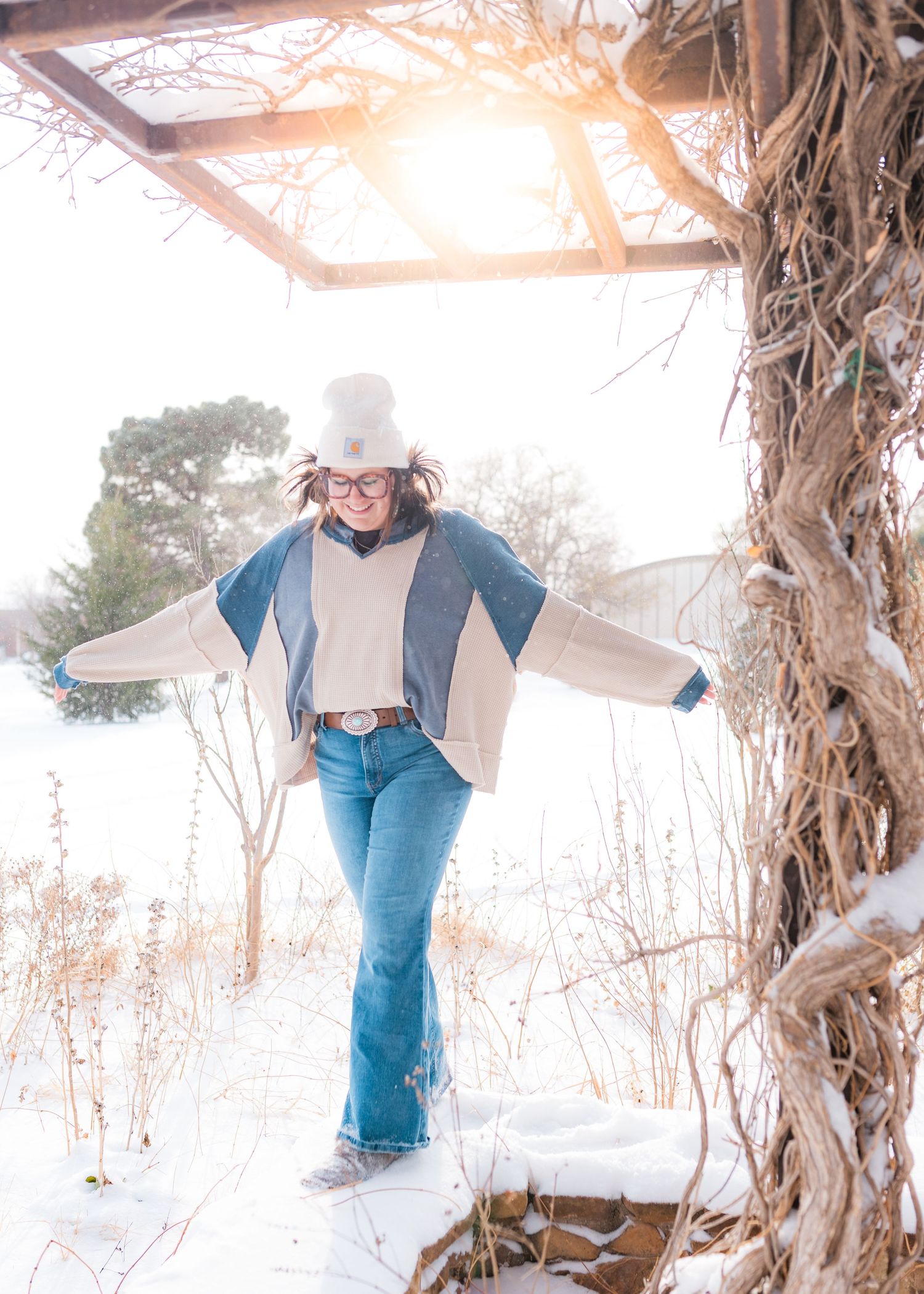 A joyful woman in flared jeans and white hat poses with arms outstretched under a snowy pergola in winter.