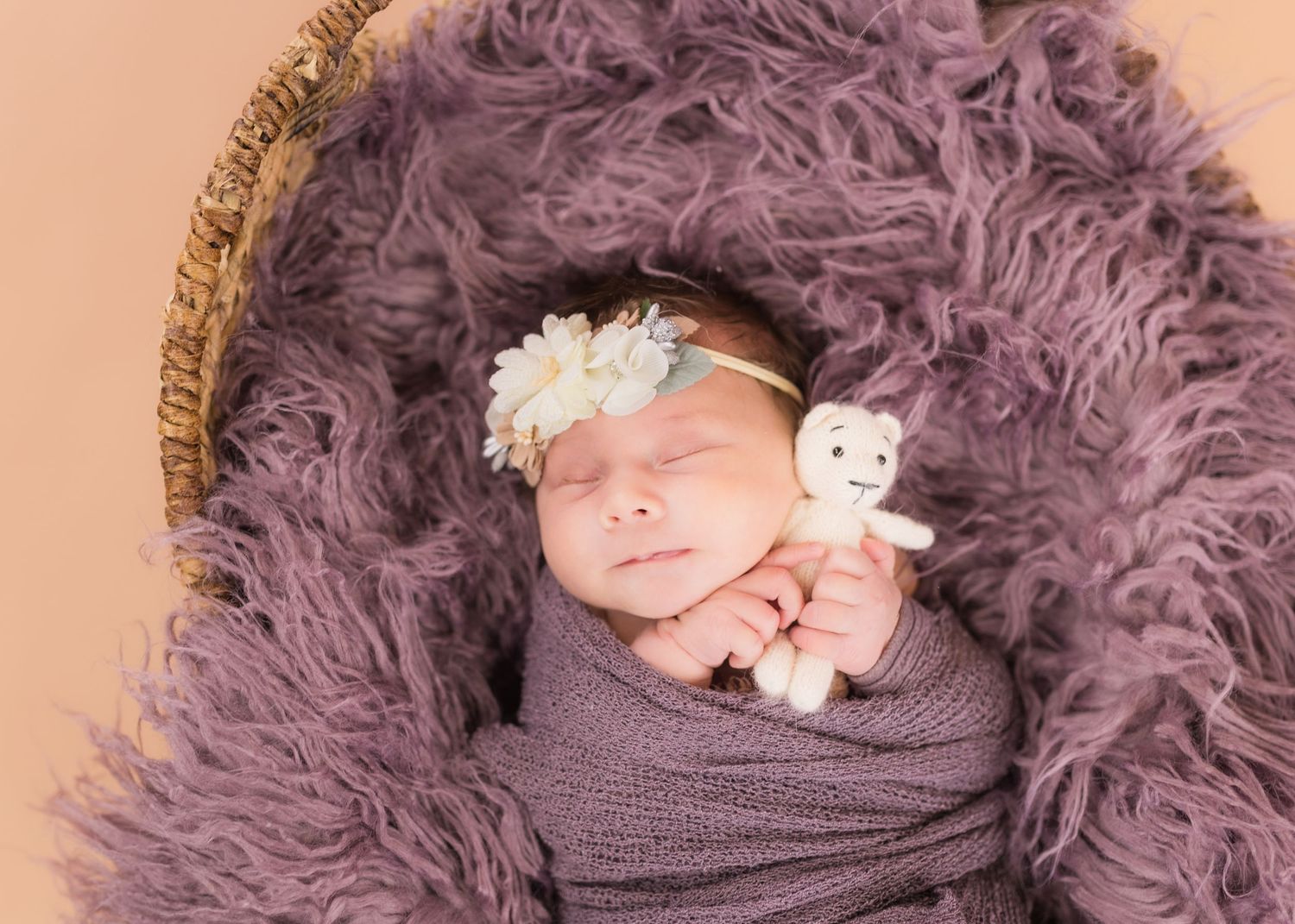 Newborn baby sleeping in a wicker basket with purple faux fur, wrapped in lavender, wearing a floral headband, holding a tiny teddy bear.