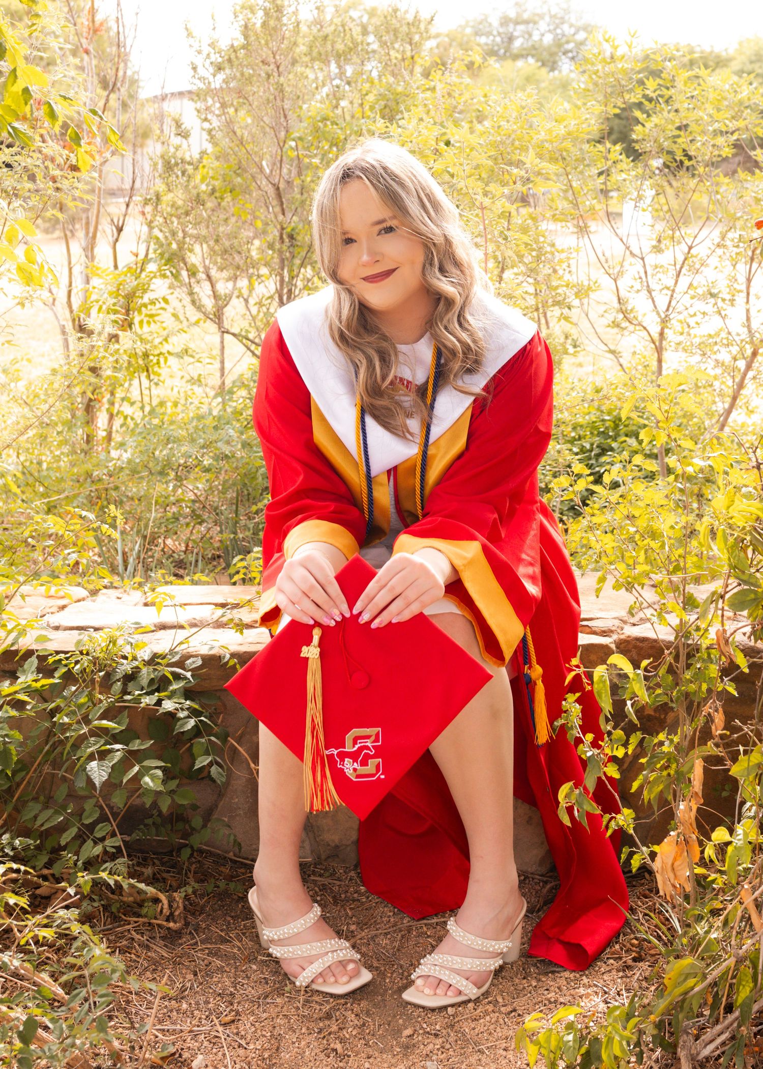 Graduate in red and gold cap and gown poses outdoors on stone wall surrounded by lush green foliage.