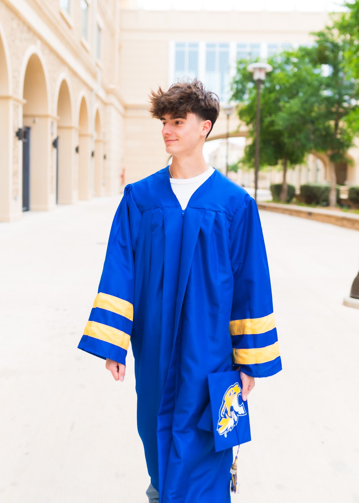 Graduate in bright royal blue cap and gown with gold stripes on sleeves stands in outdoor colonnade.
