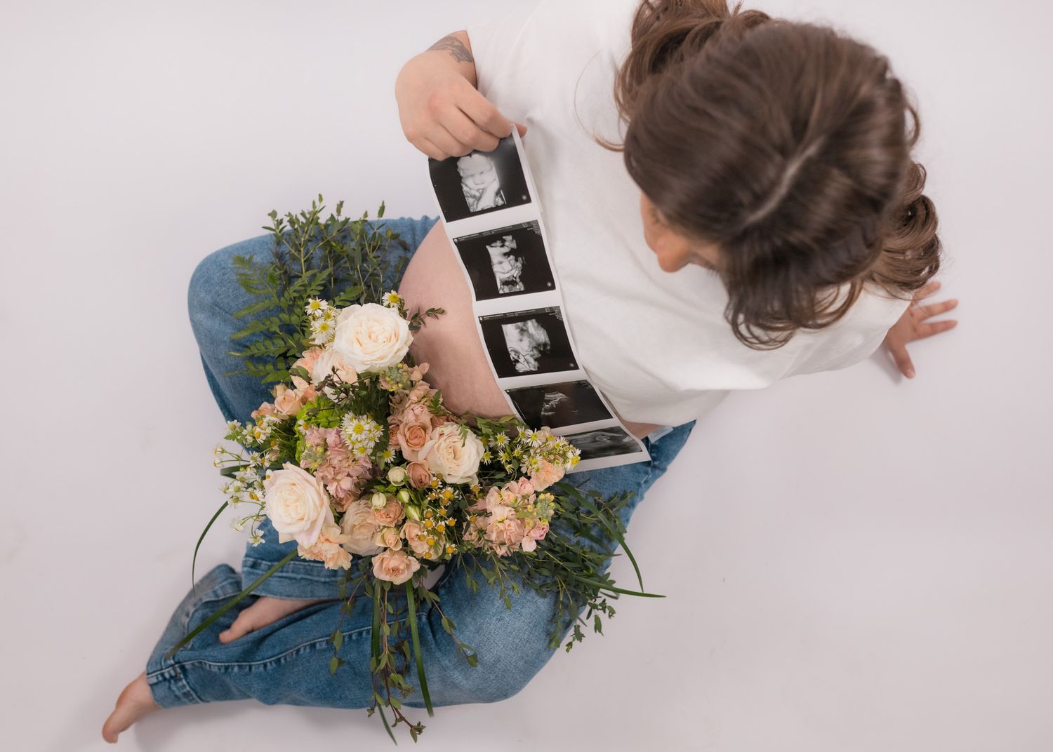 Overhead view of someone holding ultrasound photos next to a bouquet of pink roses and greenery on a blue pillow.