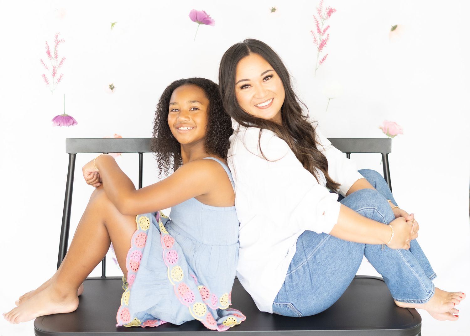 Two people in casual outfits pose back-to-back on a bench with floral wall decorations.