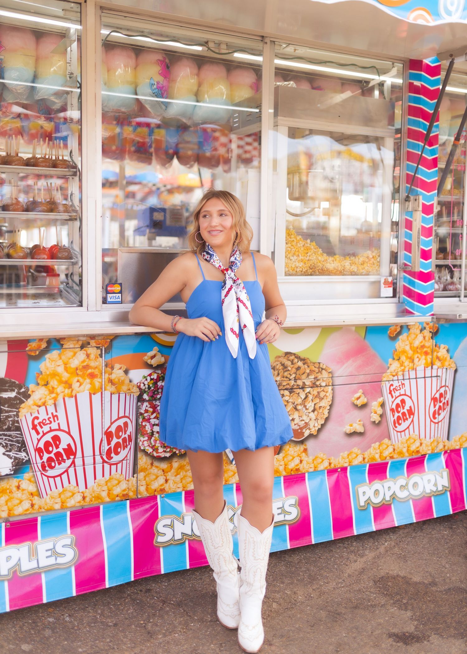Woman in blue dress and white boots posing at a colorful carnival popcorn stand.