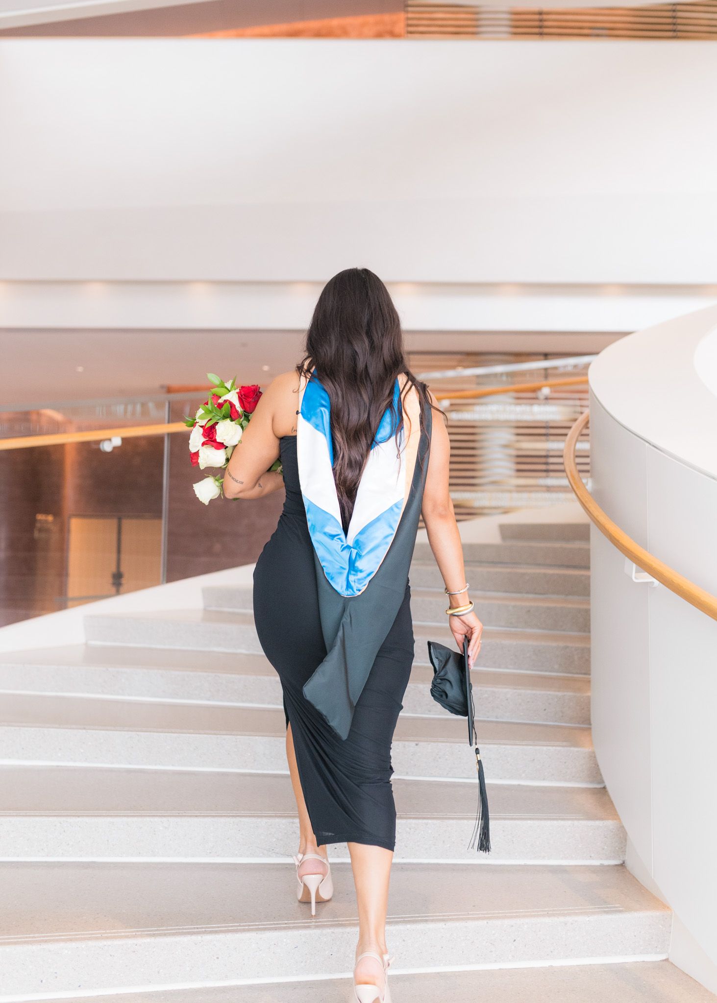 Graduate walking up modern curved stairs holding flowers and diploma.