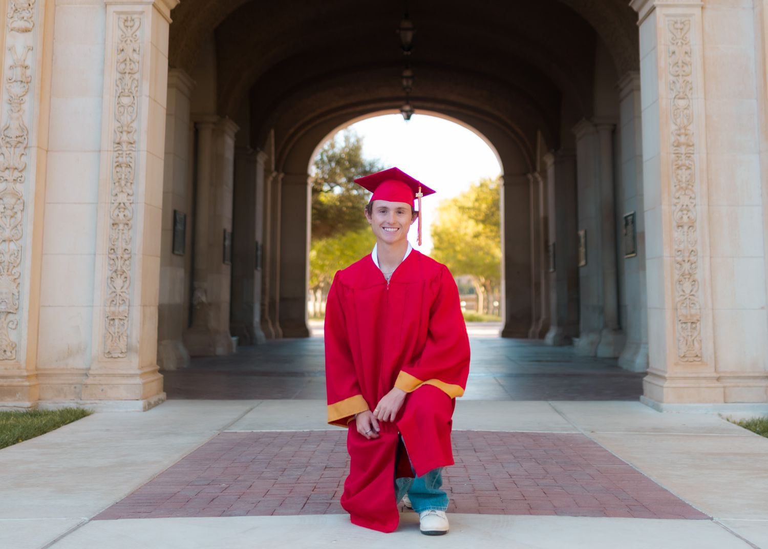 A graduate in red cap and gown kneels for a photo under a stone archway during golden hour.