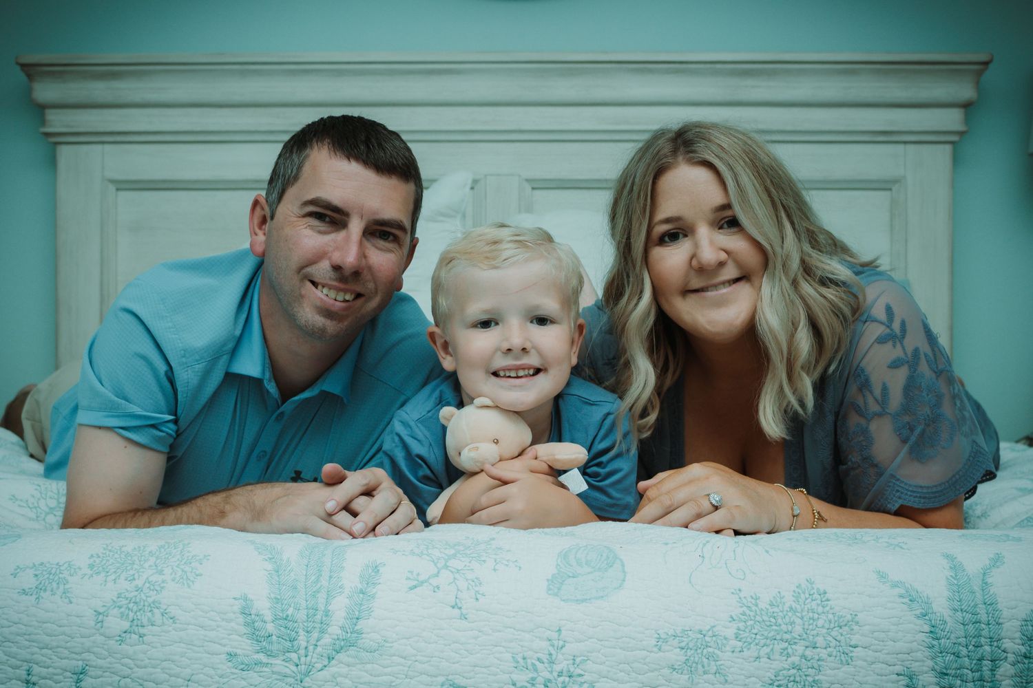 A family laying on light blue bedding captures intimate lifestyle portraits from above.