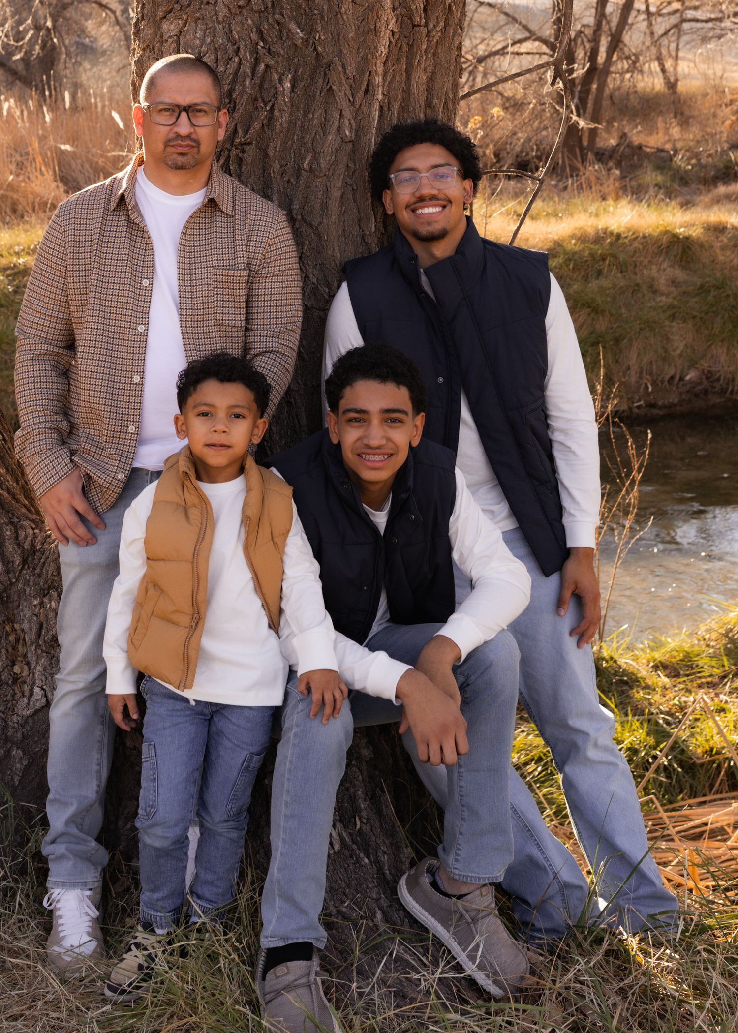 Family of four posing outdoors near a tree by a stream in autumn, wearing casual layered outfits.