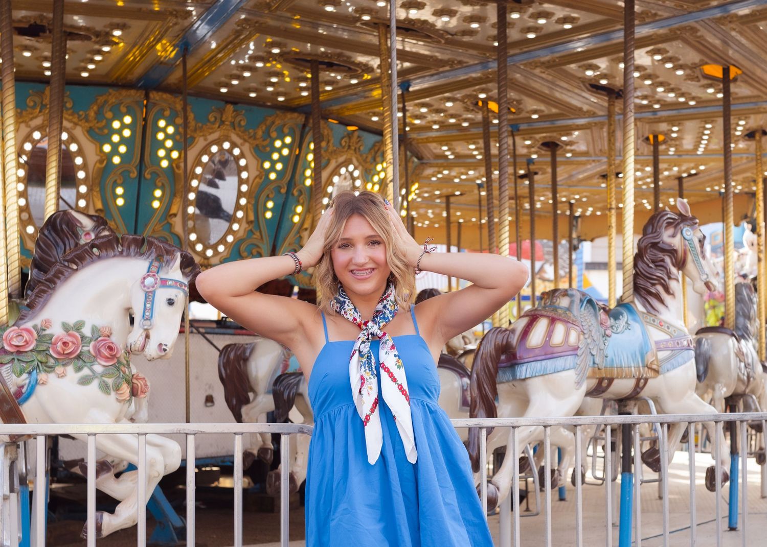Smiling blonde woman in blue dress poses in front of colorful carousel with white horses at amusement park.