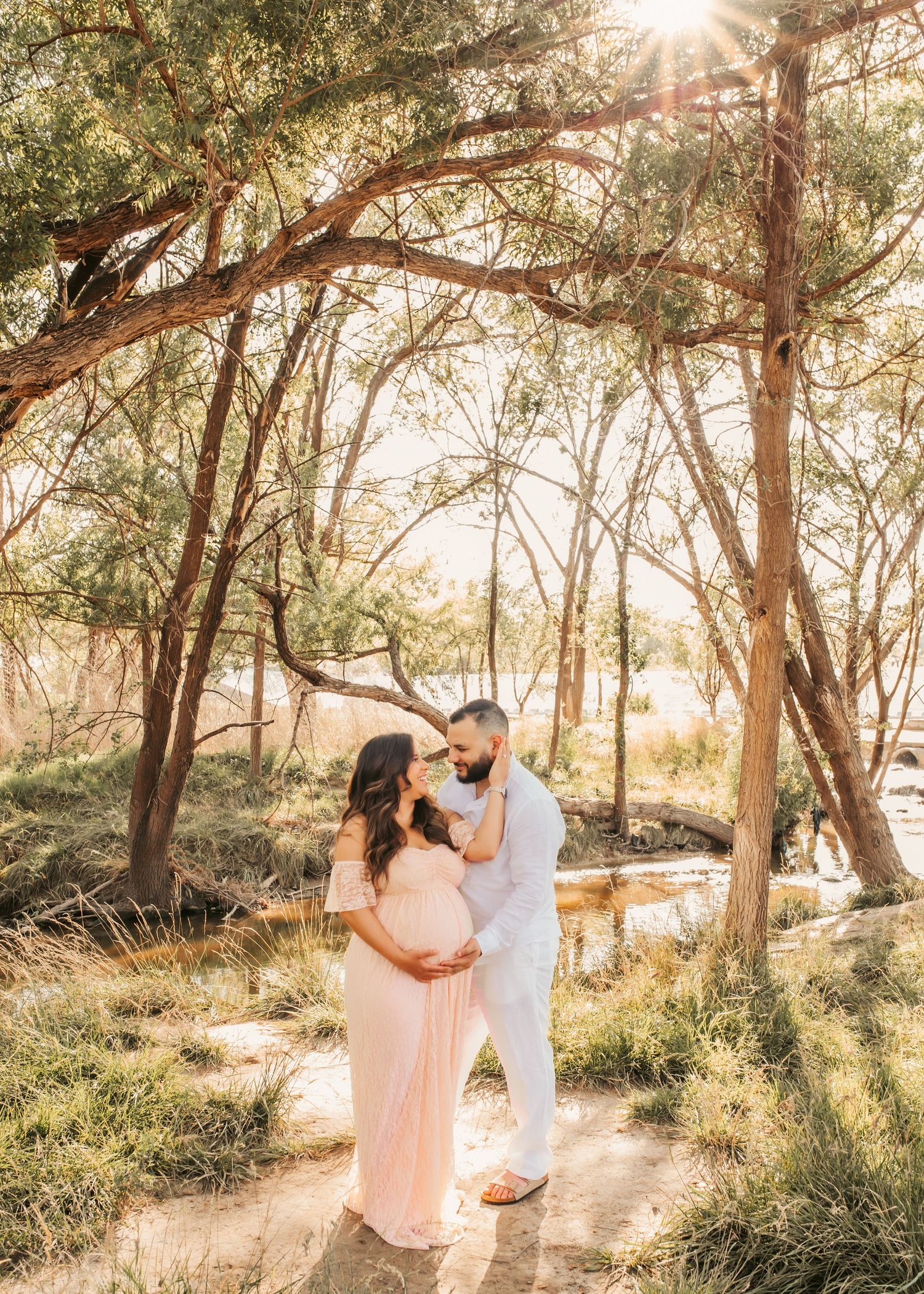 Couple sharing a tender moment in a sunlit forest, woman in pink maternity gown, man in white, surrounded by trees.