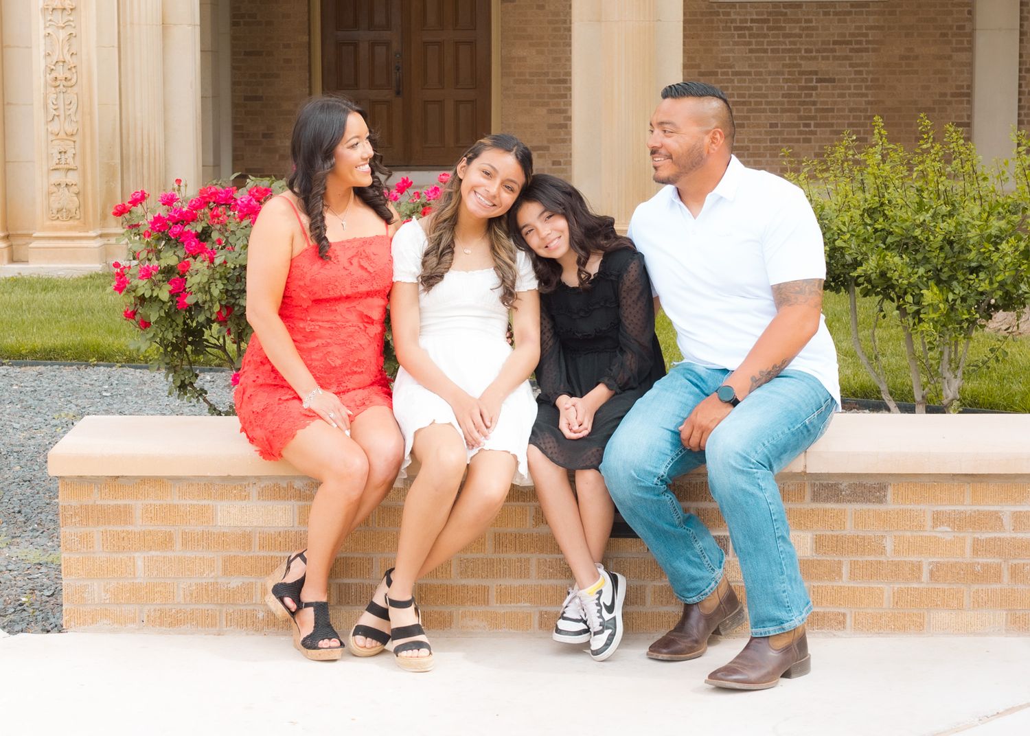 Family sitting and laughing together on a brick ledge with pink flowers in the background.