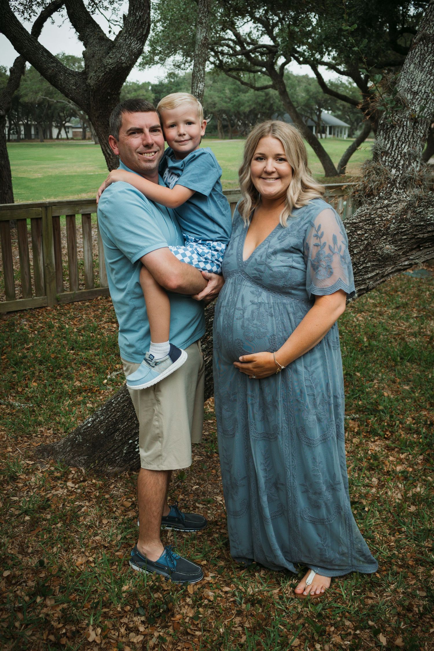 Family maternity portrait outdoors with trees and wooden fence in background.