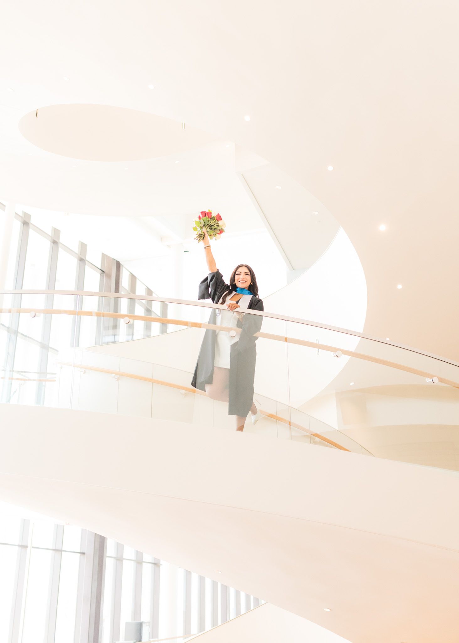 Graduate holding flowers poses on curved modern staircase in bright interior.