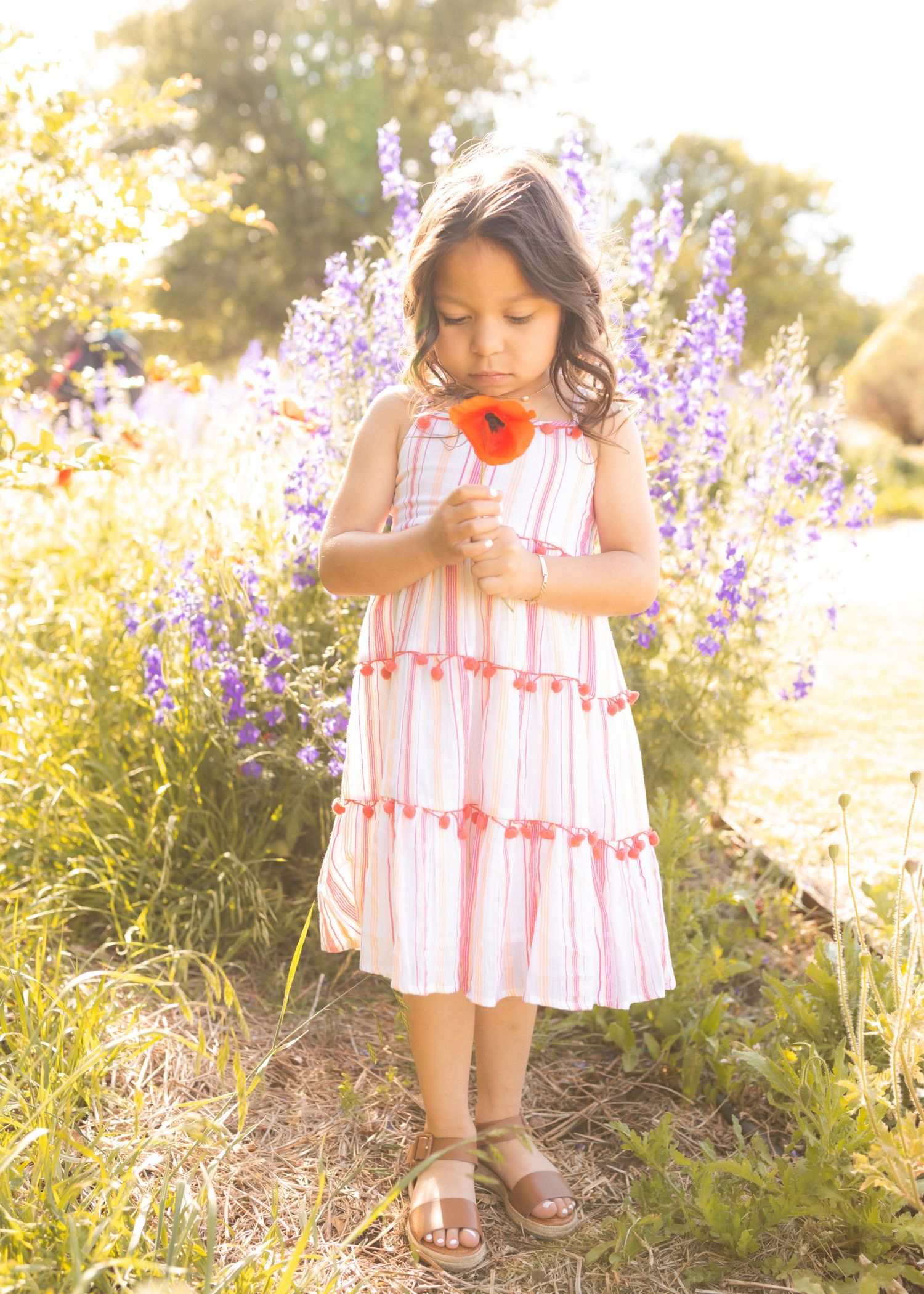 Young girl in striped dress smelling red flower in purple wildflower field.