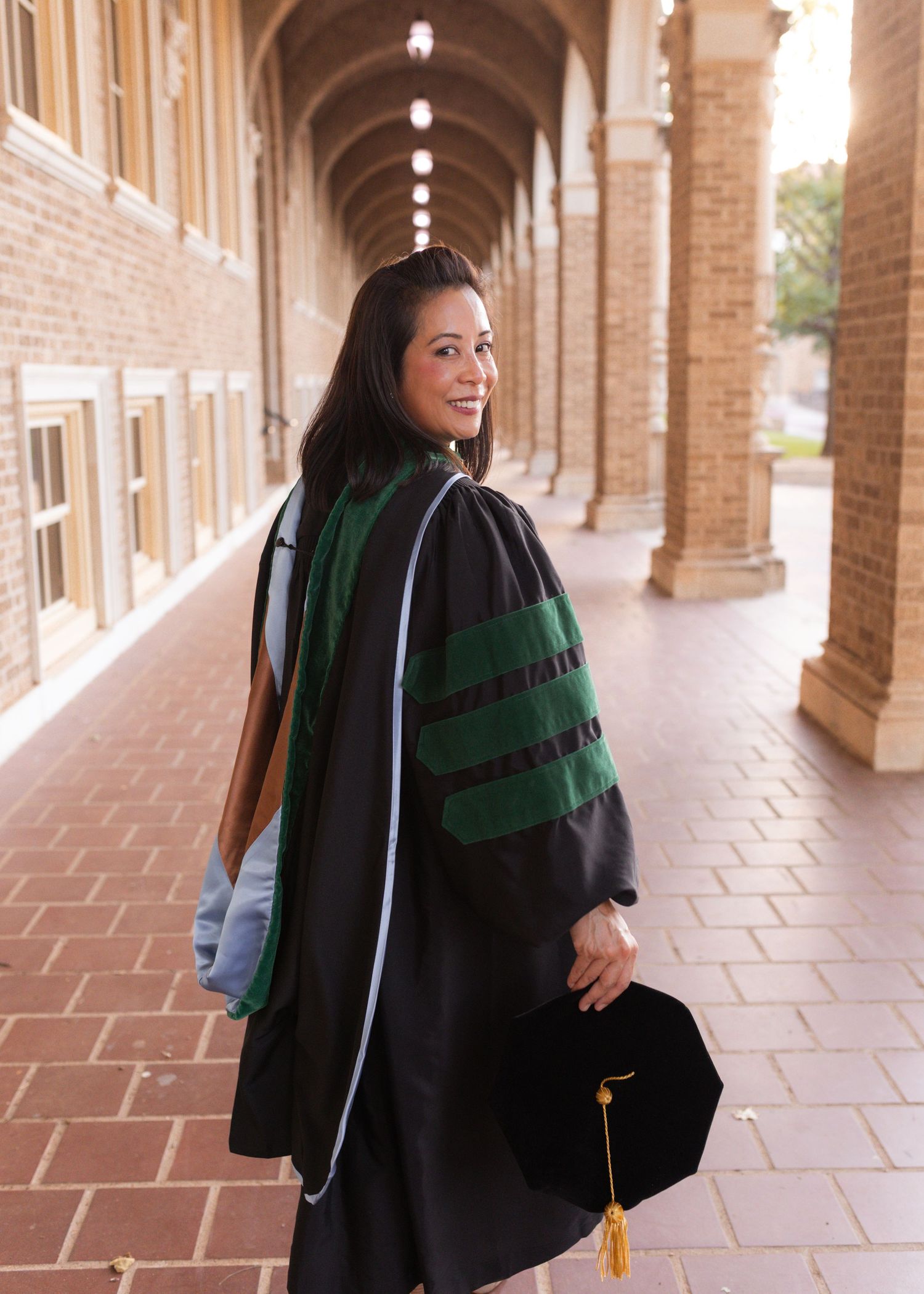 Woman in black doctoral graduation gown with green velvet stripes holds cap in arched brick hallway.