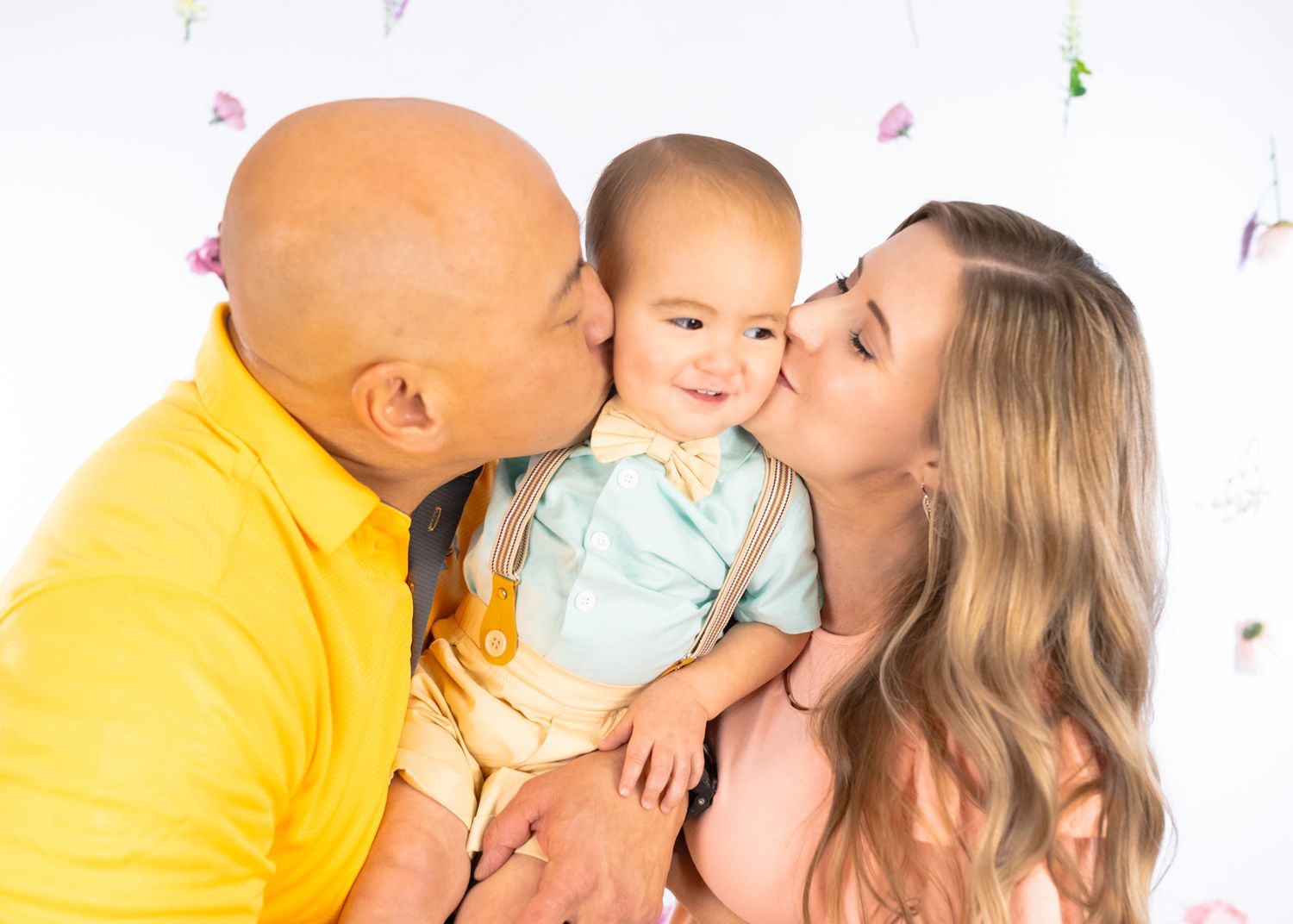 Parents share kisses with their baby against a delicate floral backdrop in a studio setting.