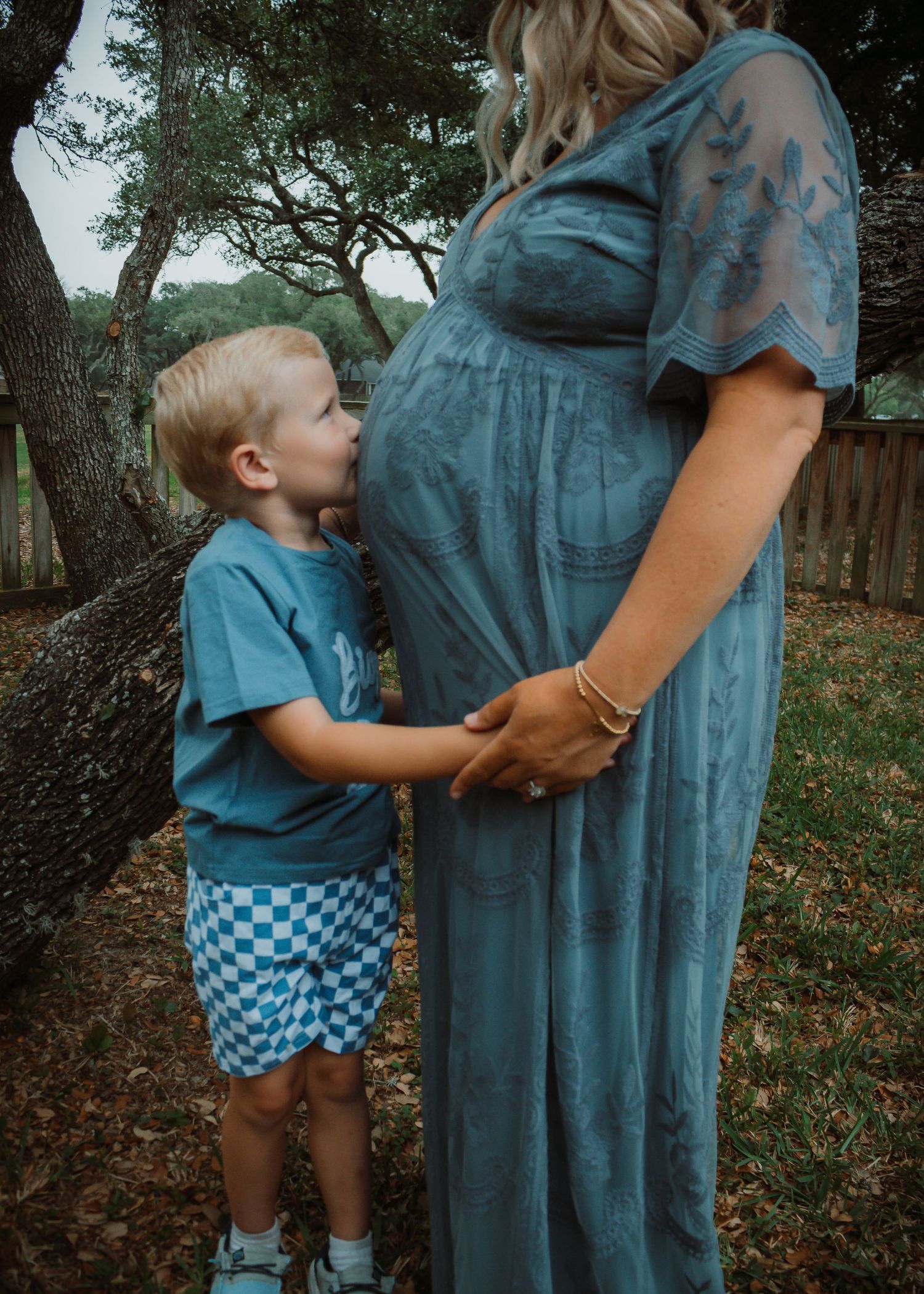 Young child in blue shirt touching pregnant belly in outdoor setting.