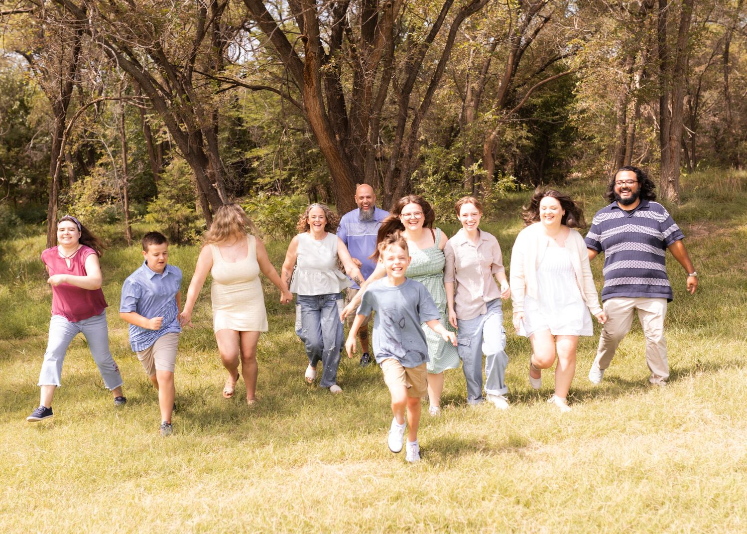 Large family group joyfully running through a sunny green meadow surrounded by tall trees on a warm day.