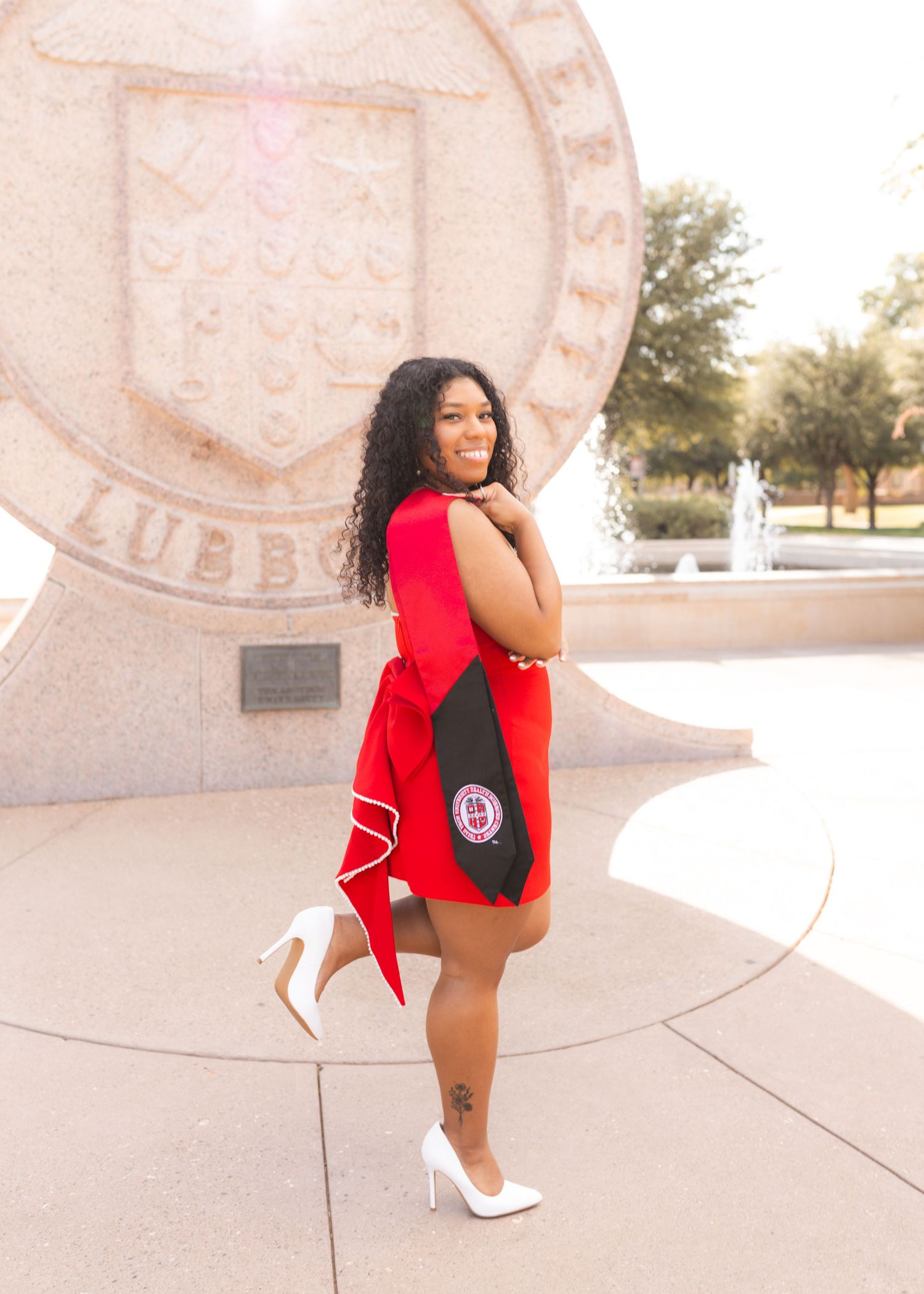 Woman in red graduation attire poses confidently in white heels near a university seal monument outdoors.