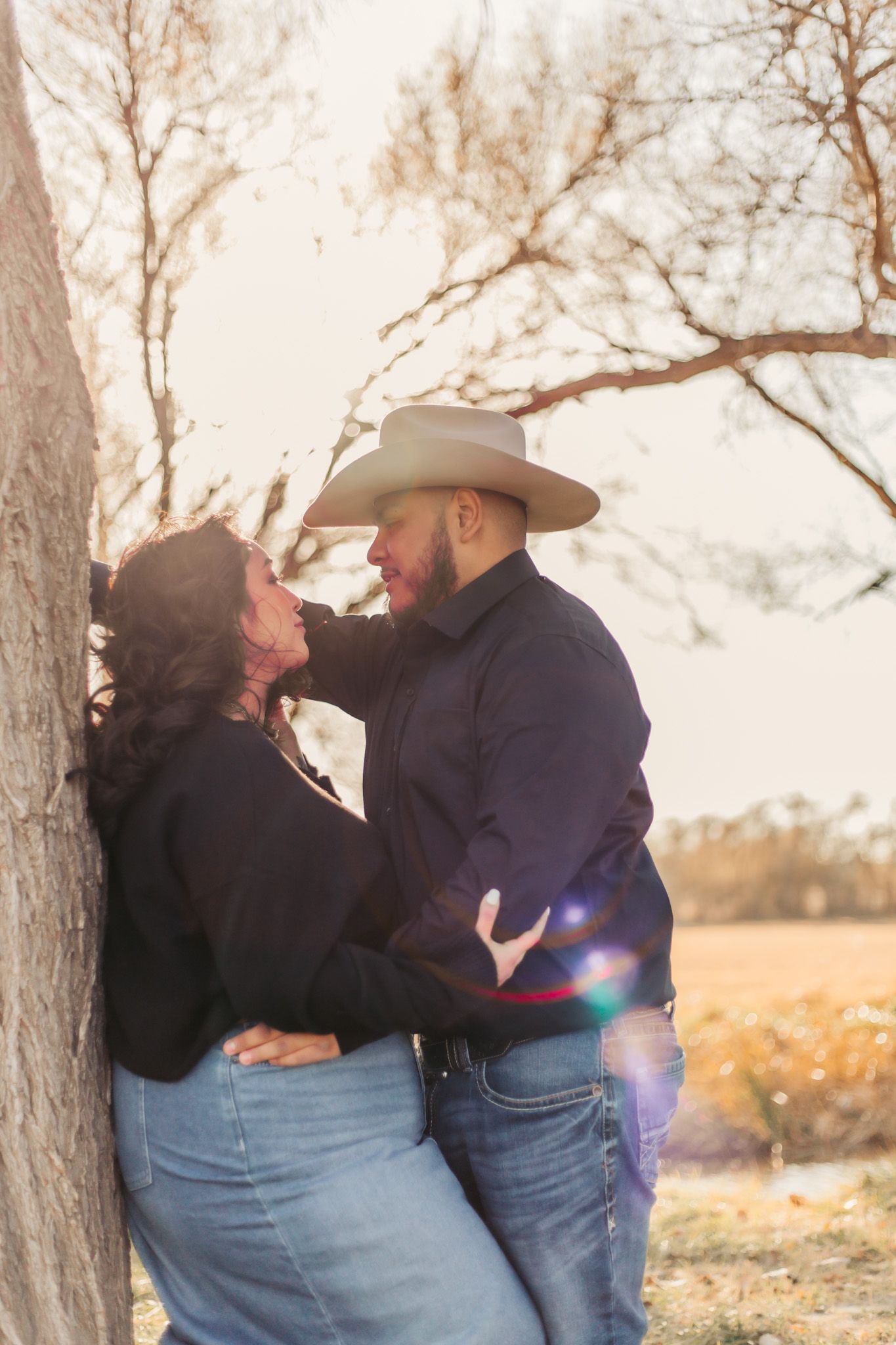 Couple in western wear sharing romantic moment against tree during sunset photoshoot.