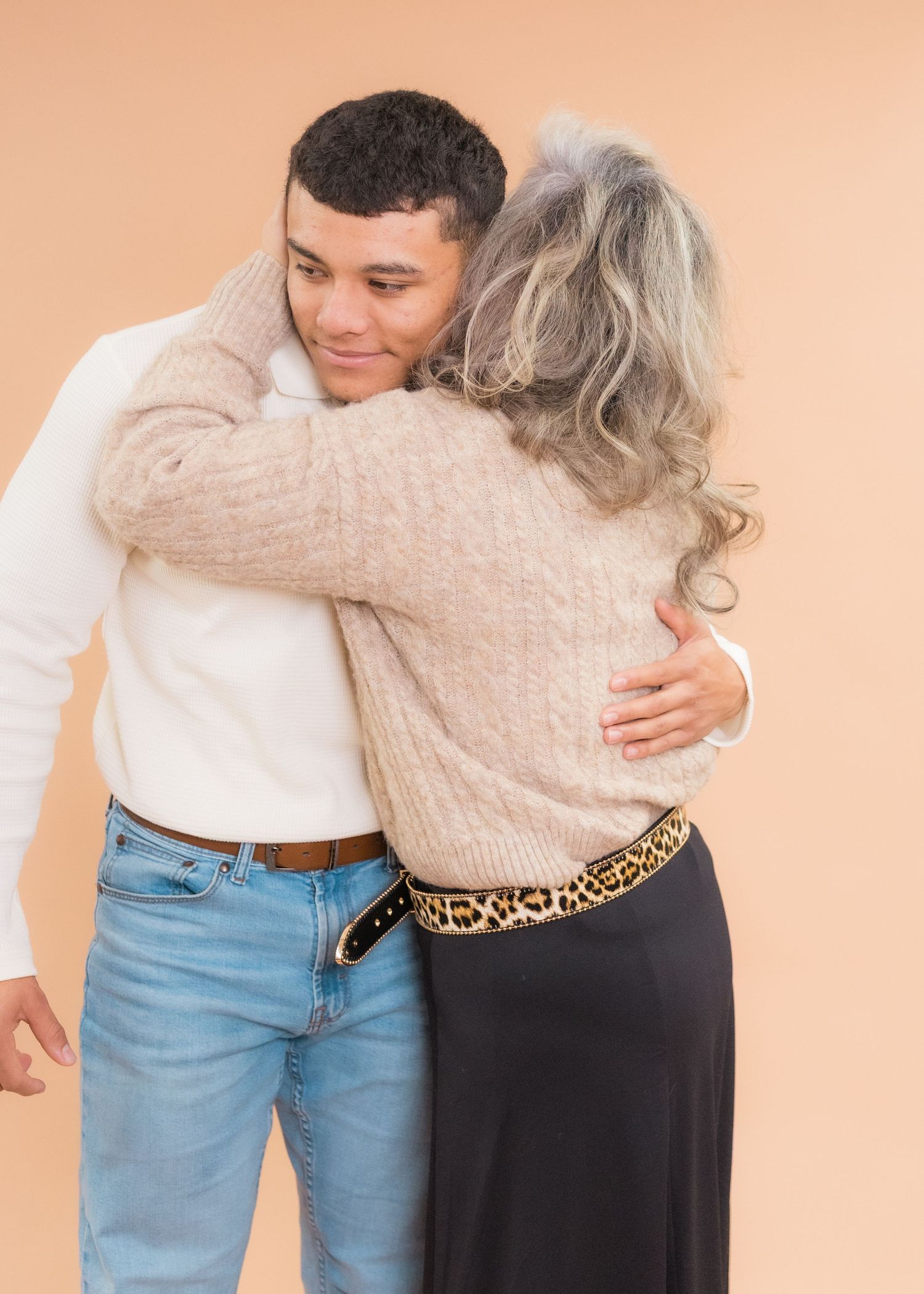 A young man and older woman share a warm embrace, both wearing cozy sweaters against a beige background.