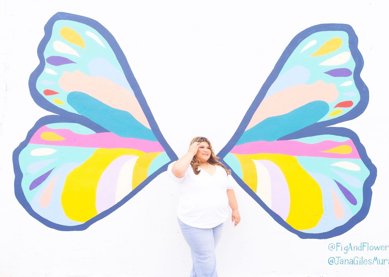 Woman posing in front of colorful butterfly wings mural on white wall, wearing white top and light jeans.