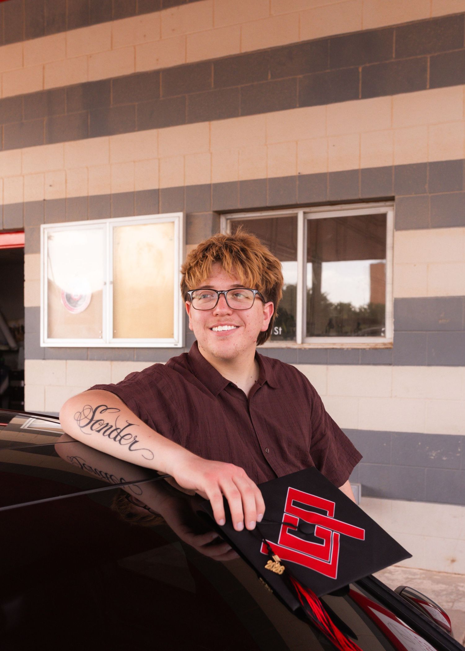 Smiling graduate leaning on car with graduation cap and Senior tattoo on arm, wearing maroon shirt.