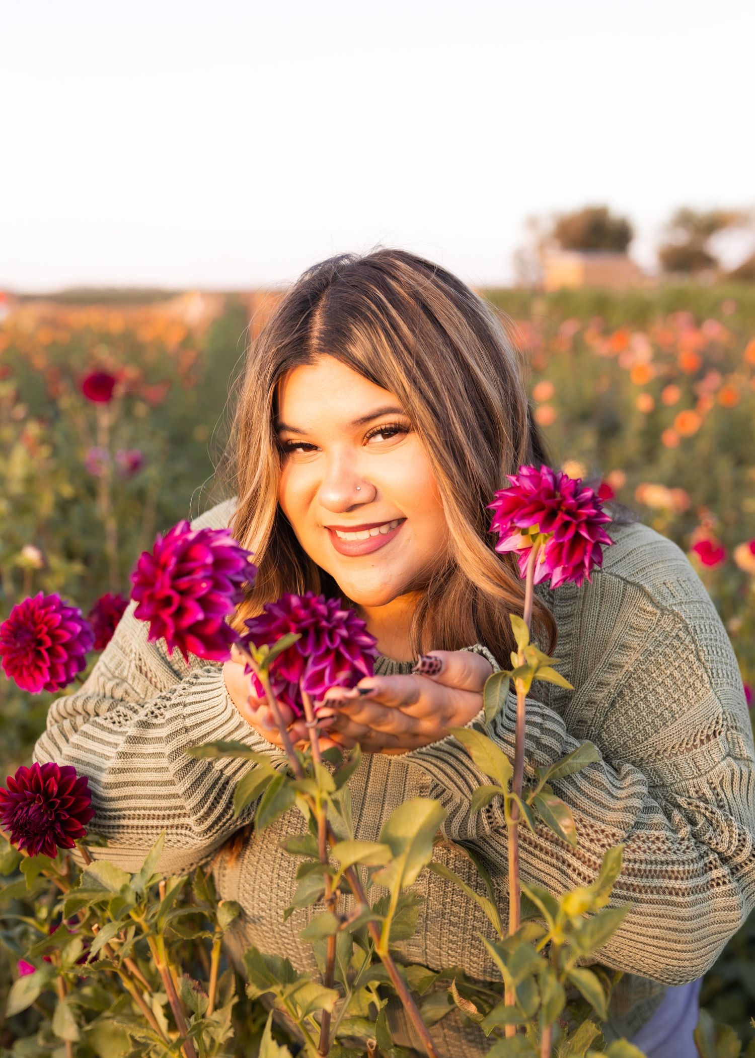 Young woman smiling in a vibrant flower field, holding pink dahlias, wearing a sage green knit sweater at golden hour.