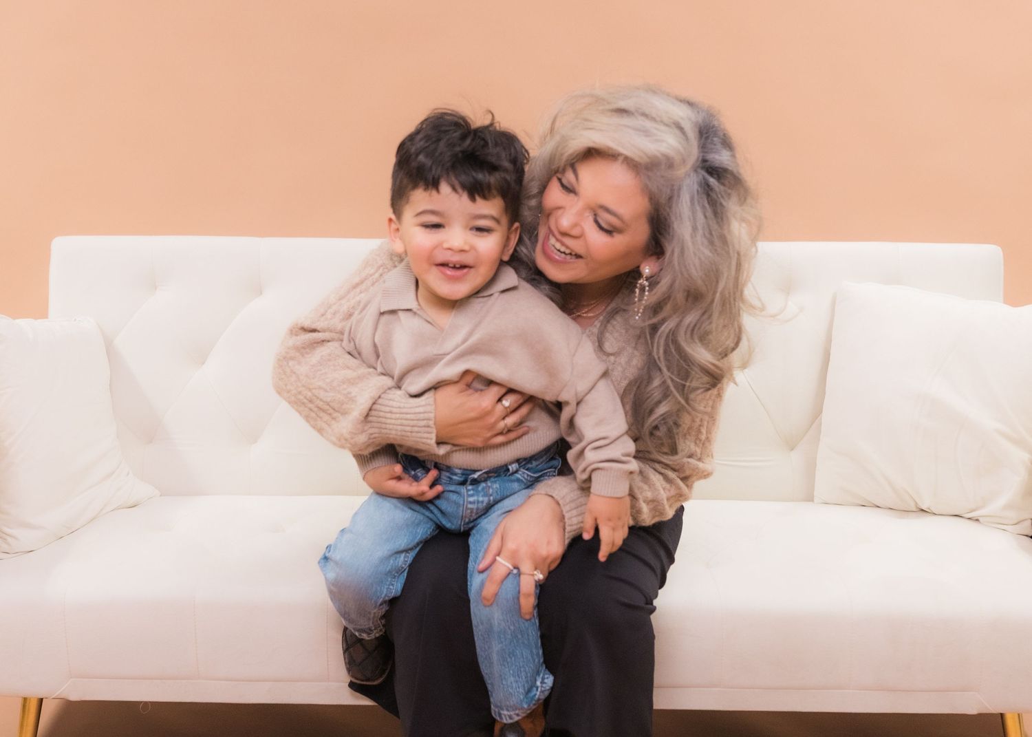 Grandmother with gray hair hugging a smiling toddler boy on a white couch against a beige background.