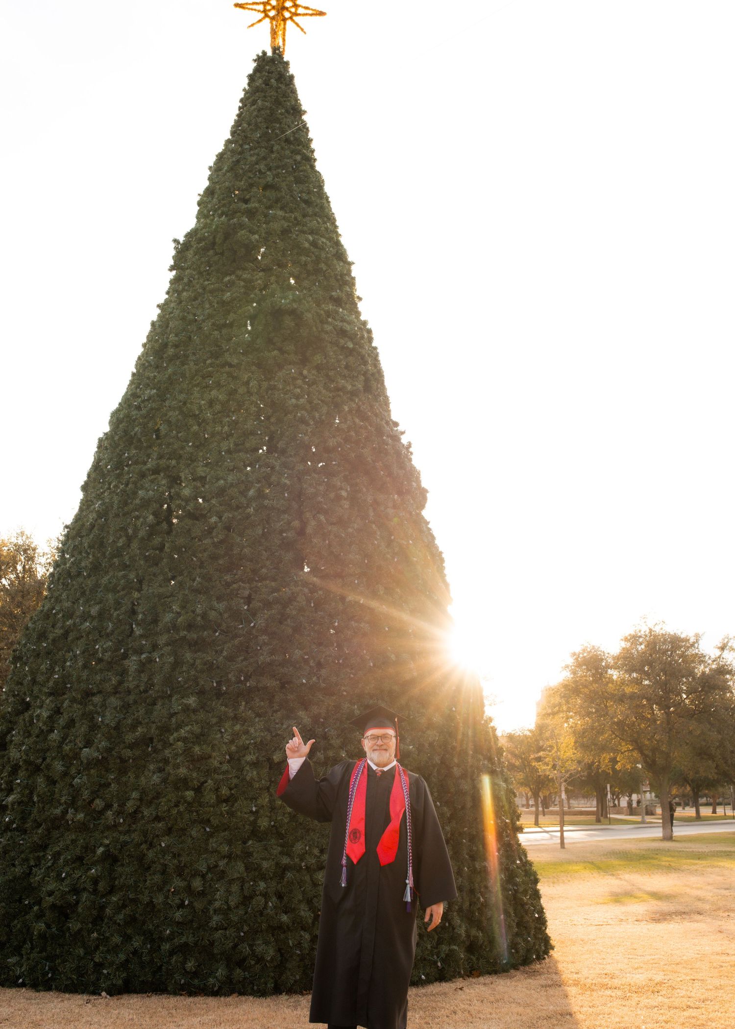Graduate in cap and gown poses proudly in front of large Christmas tree with glowing star topper at sunset.