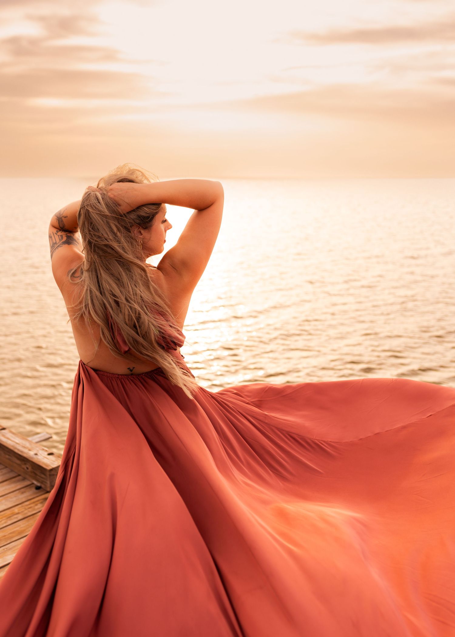 Woman in flowing terracotta gown poses on dock at golden sunset overlooking calm ocean waters.
