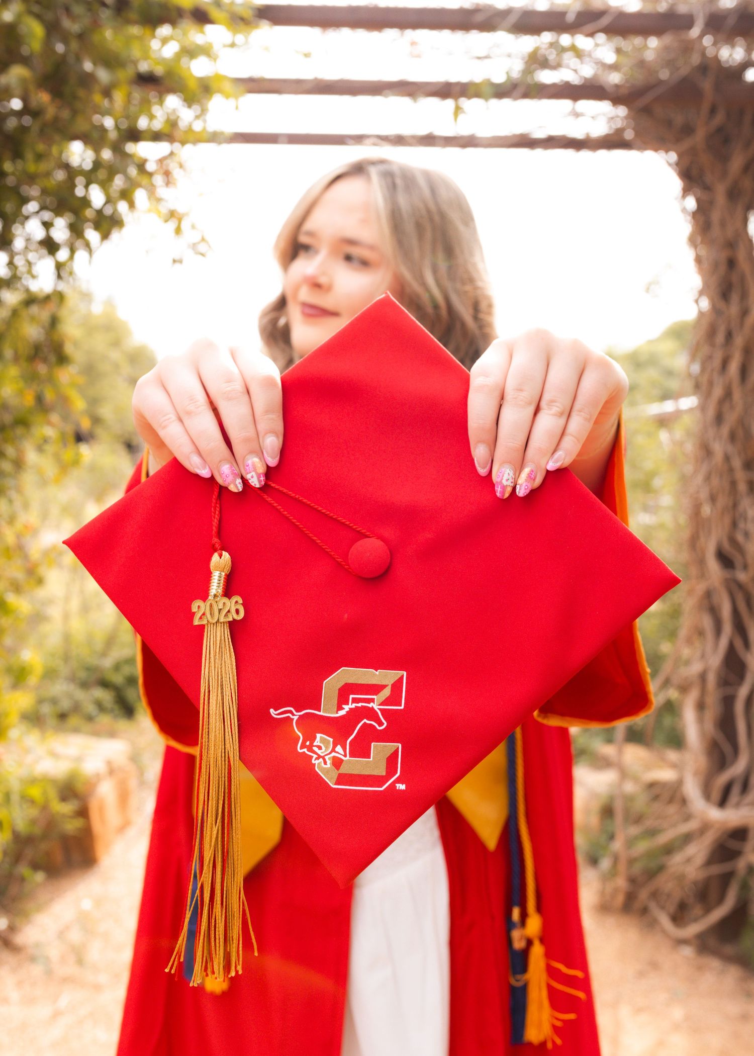 Graduate holds red mortarboard cap with gold tassel and university logo, wearing graduation gown outdoors.