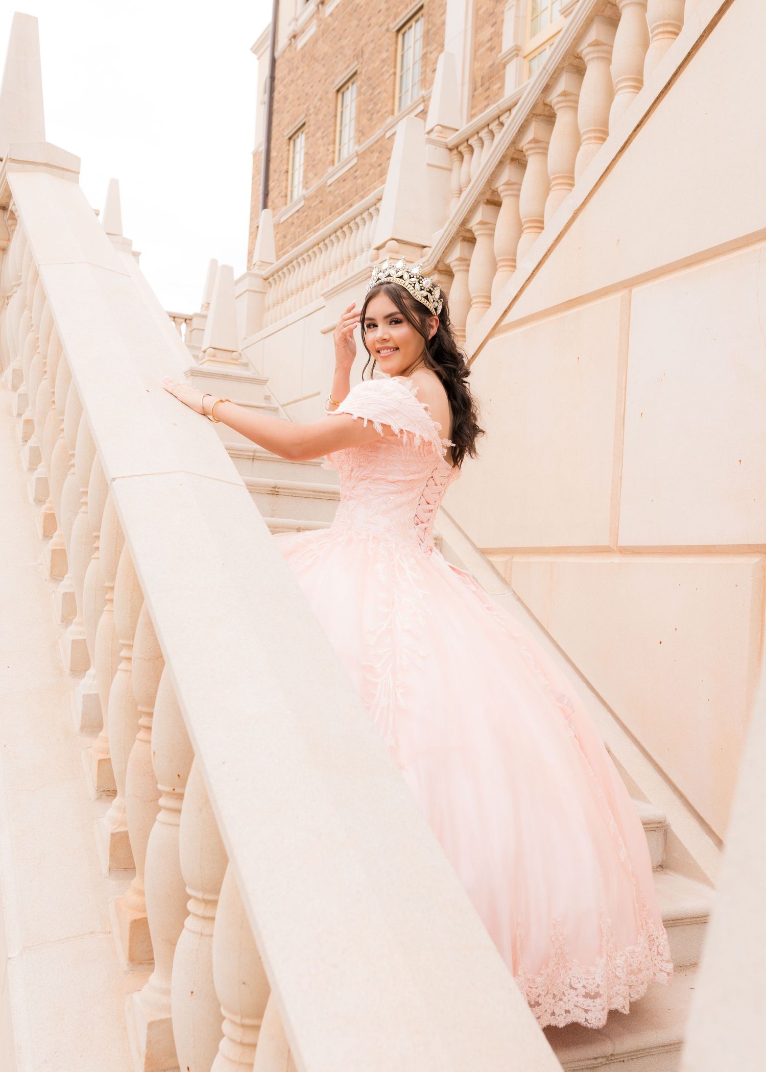 Young woman in pink quinceañera ball gown and crown poses gracefully on white balustrade staircase.