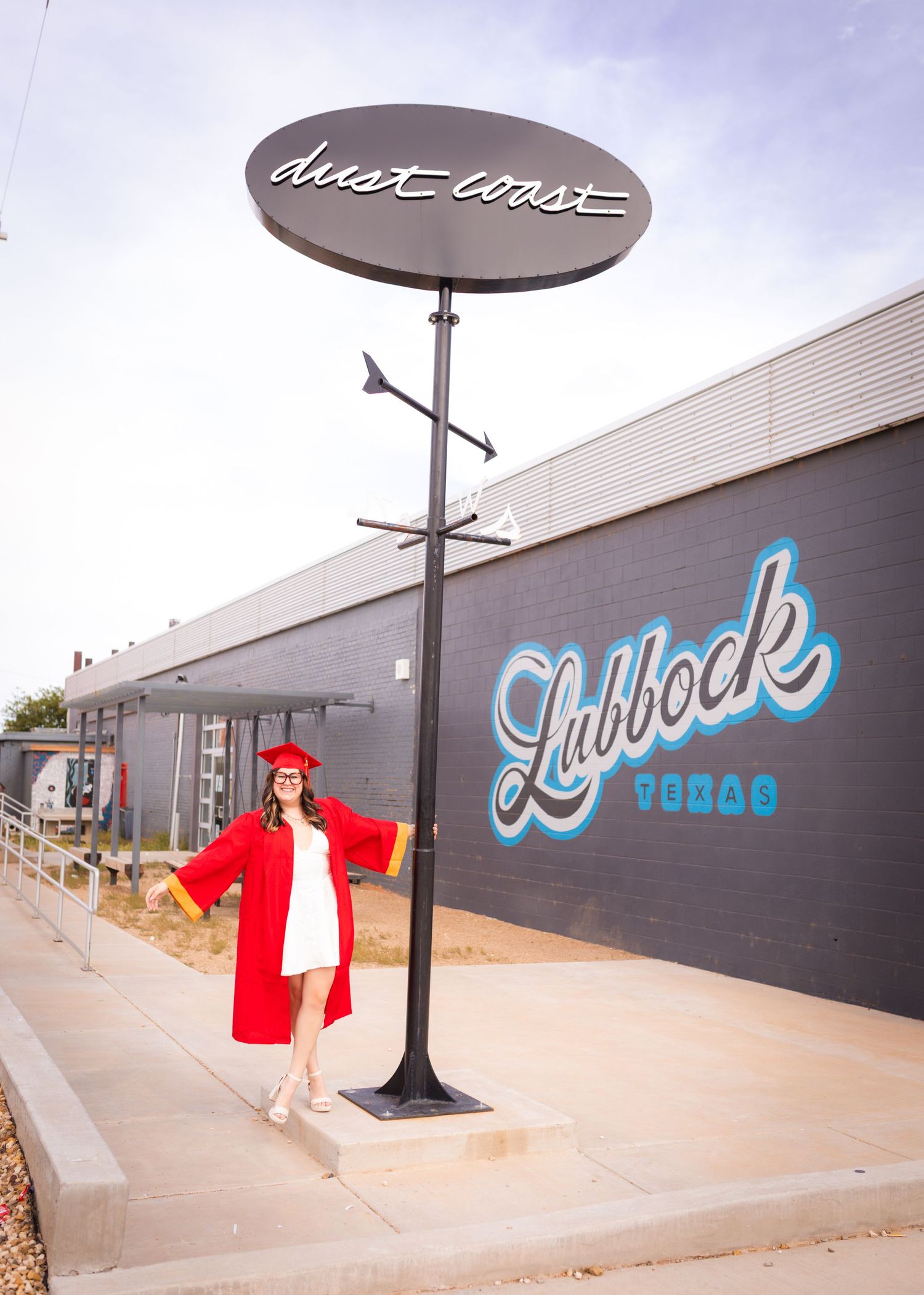 Graduate in red cap and gown poses by Dust Coast sign with Lubbock Texas mural in background.