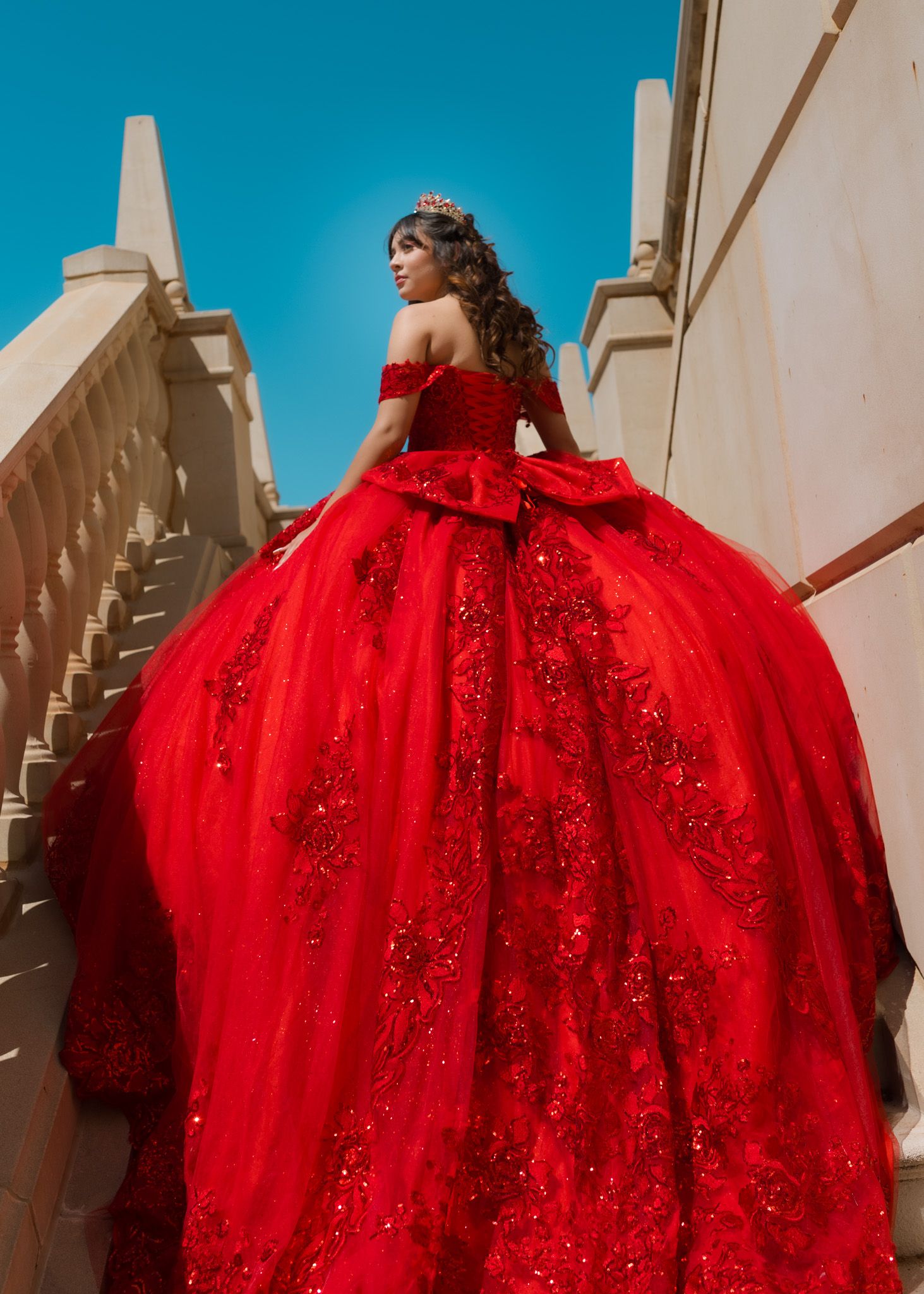 A stunning red ball gown with sparkly details and off-shoulder design photographed on an outdoor staircase against blue sky.