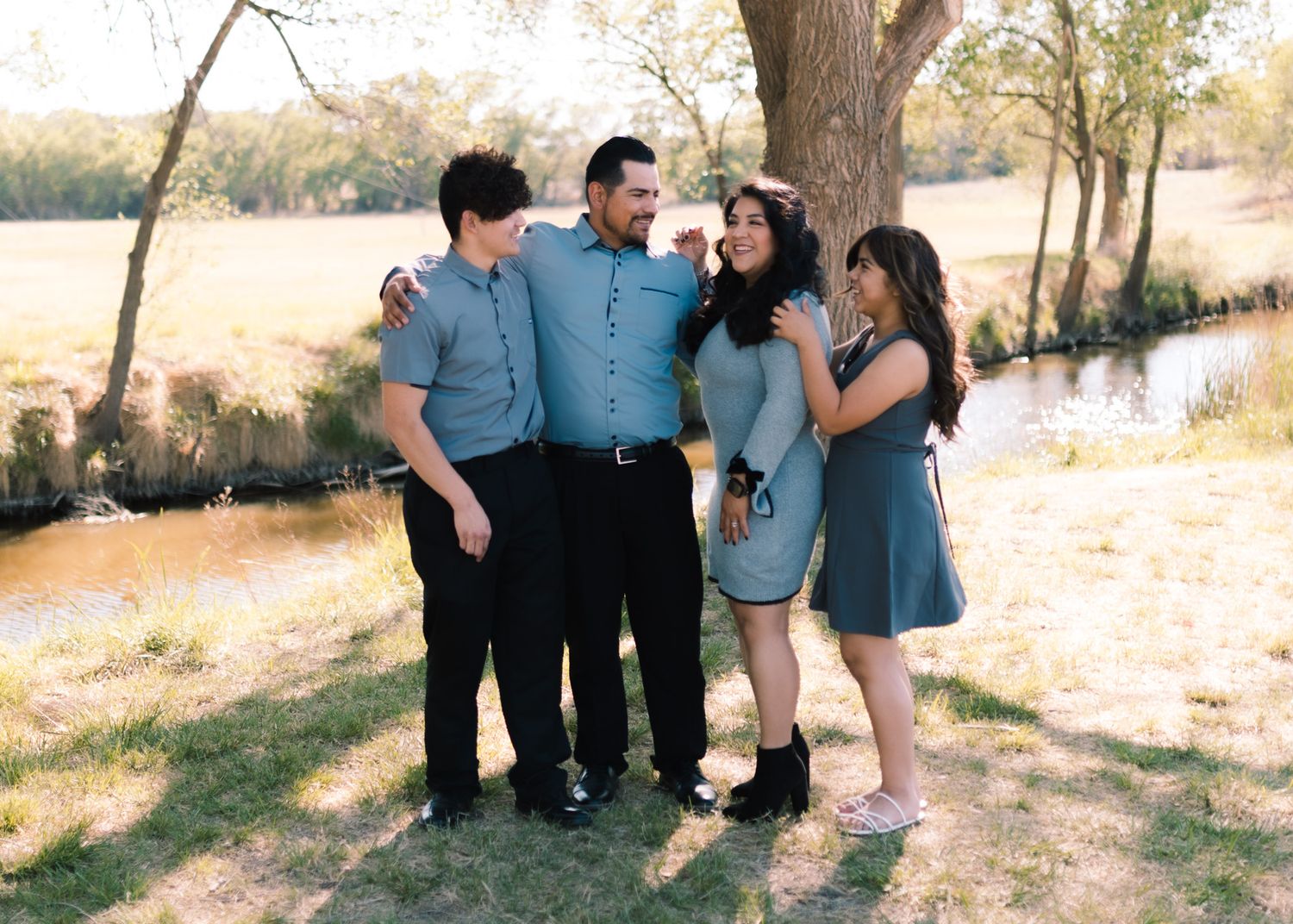 A cheerful group in coordinating blue outfits poses together under a tree near a scenic lake on a sunny day.