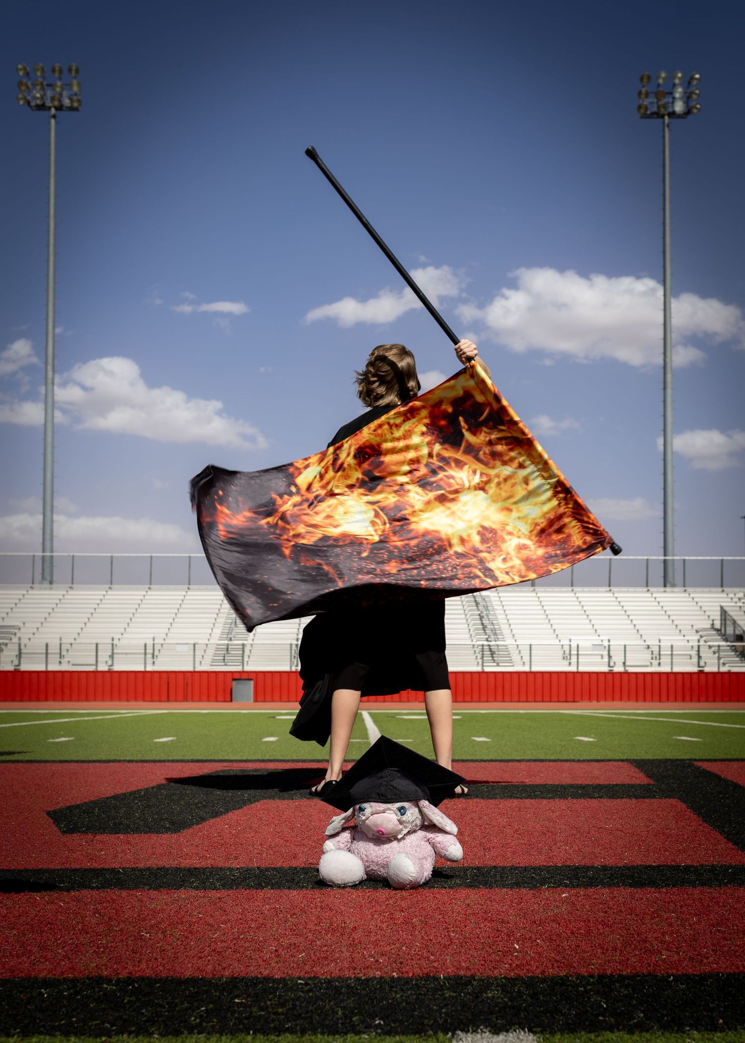 A color guard performer waves a flame-printed flag on a red track field with stadium lights against a blue sky.
