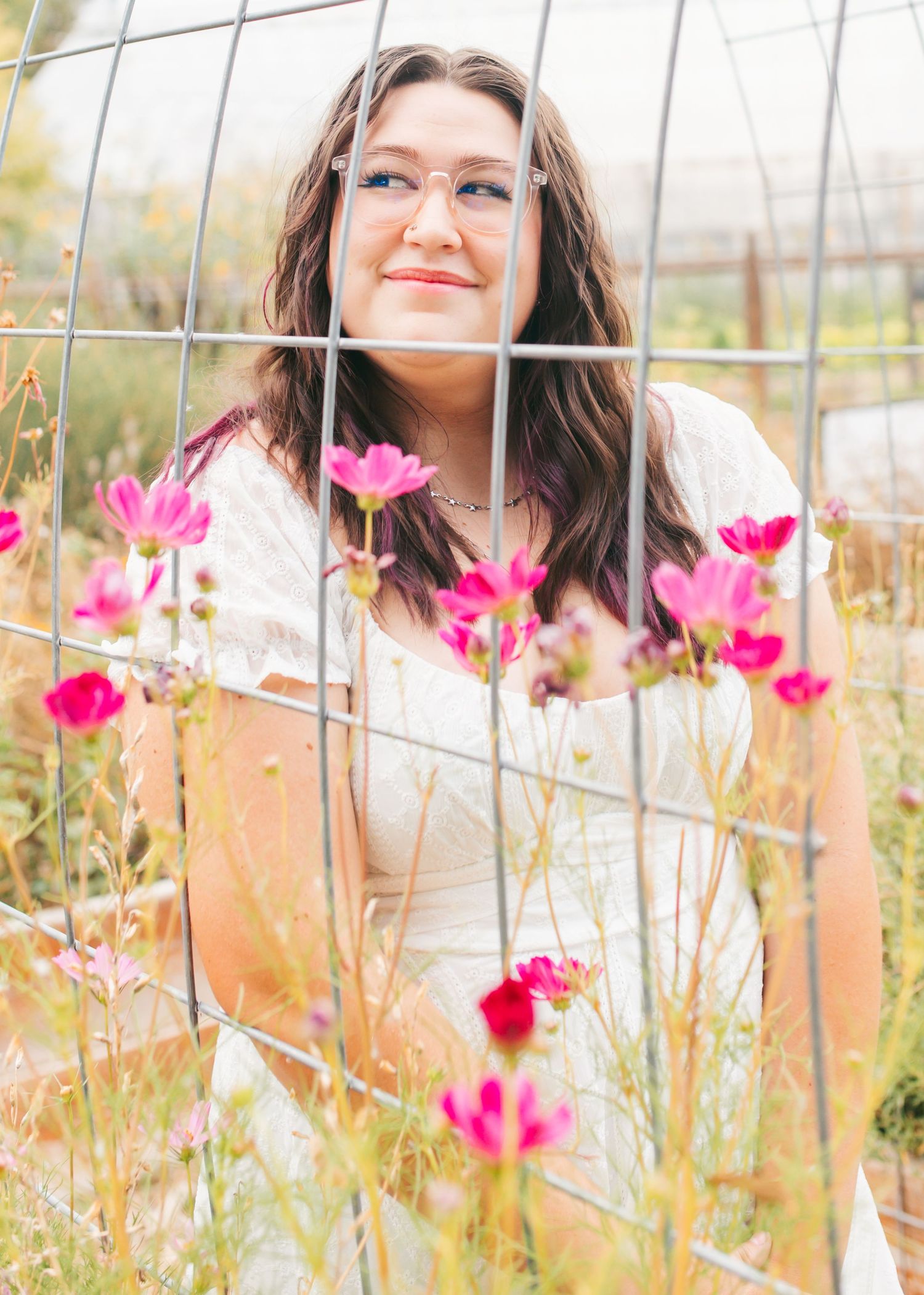Young woman with glasses poses among pink cosmos flowers behind a wire garden trellis in a white dress.