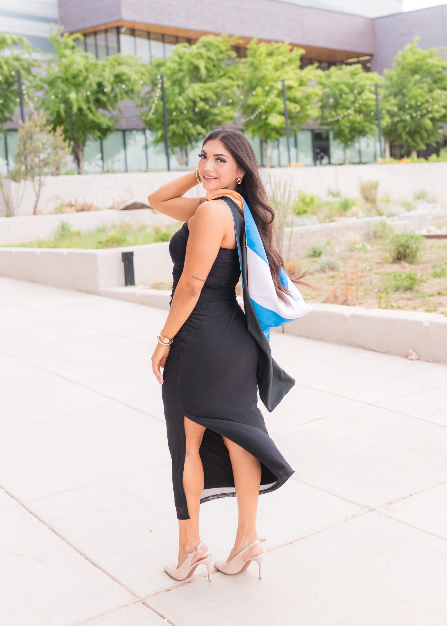Woman in fitted black dress with side slit poses outdoors near modern building.