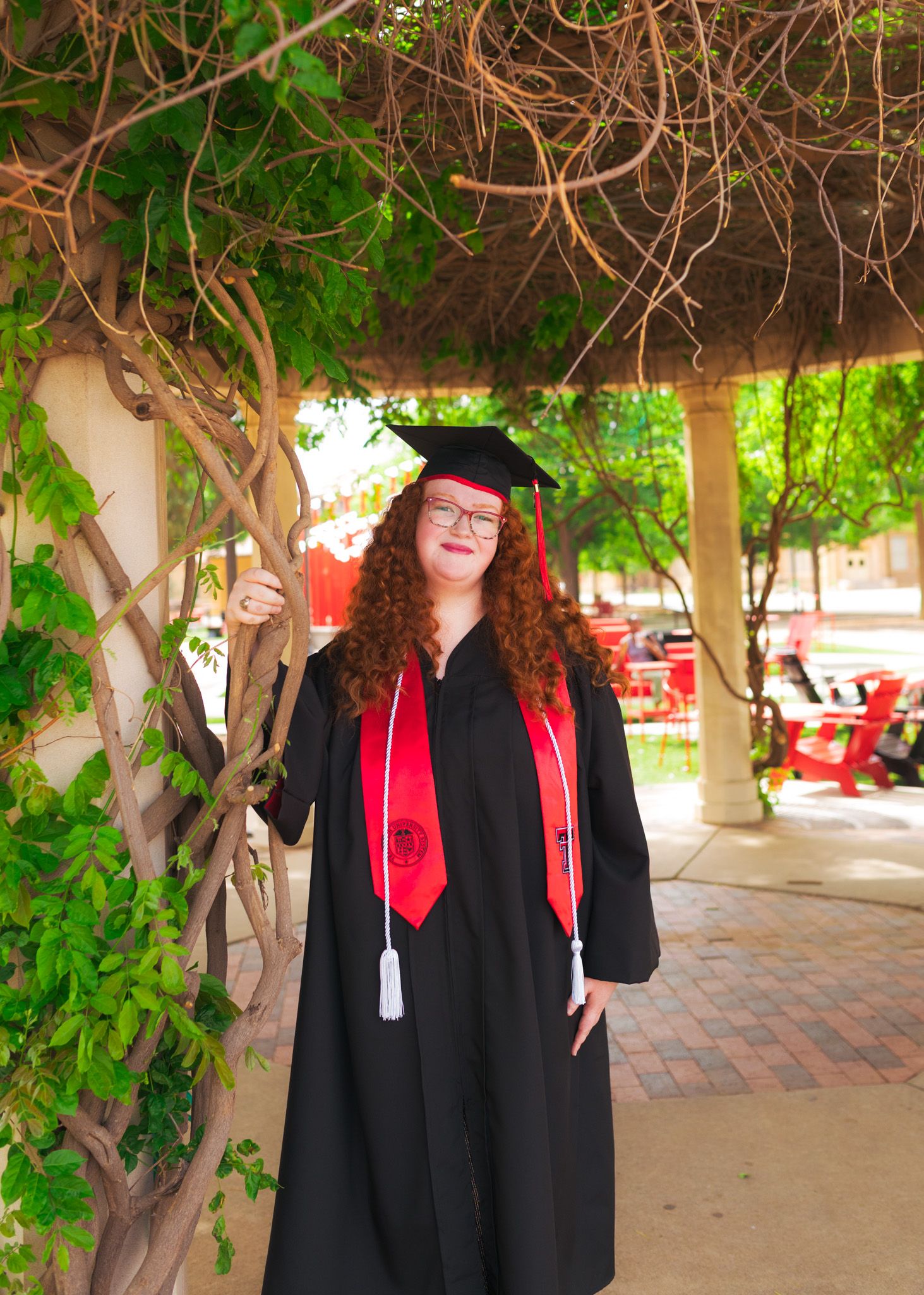 A graduate wearing academic regalia with red stole poses under vine-covered pergola.