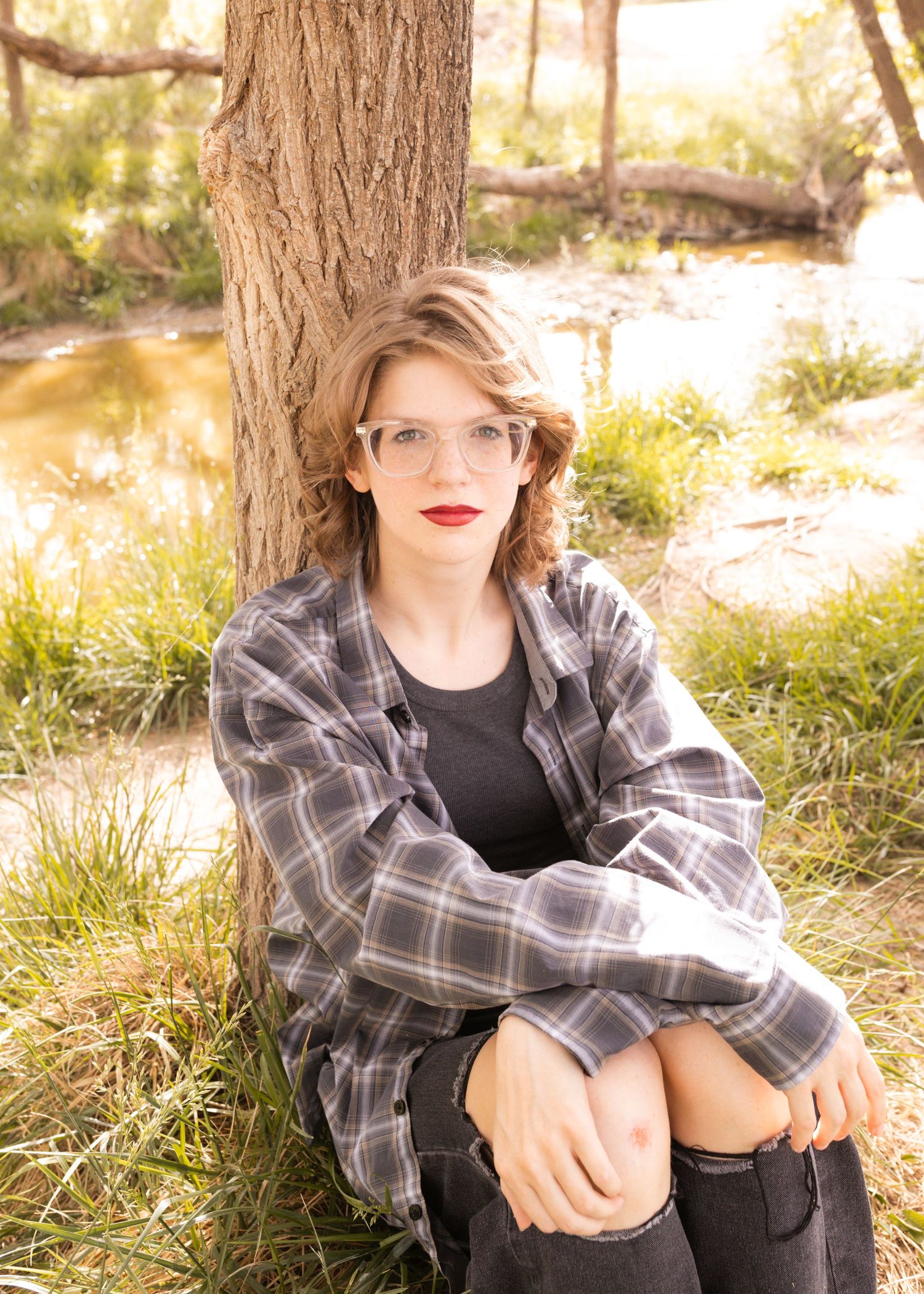 Young woman with curly hair and glasses in plaid shirt sits against tree by stream in natural outdoor setting.
